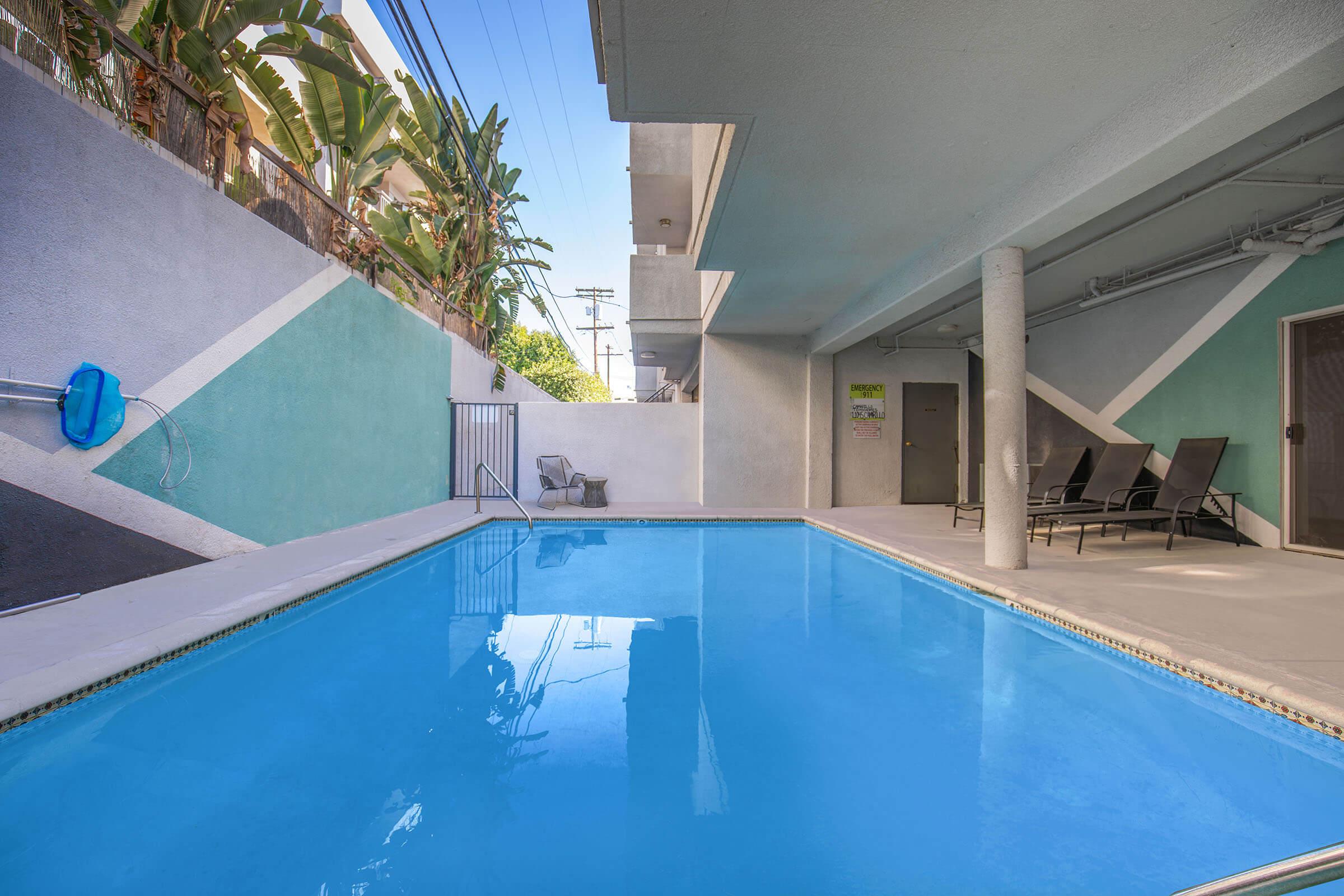 A tranquil swimming pool surrounded by a modern, geometric design on the walls. The pool reflects the clear blue sky, with a few lounge chairs positioned nearby. Green tropical plants add a lively touch to the scene, creating a relaxing outdoor space.