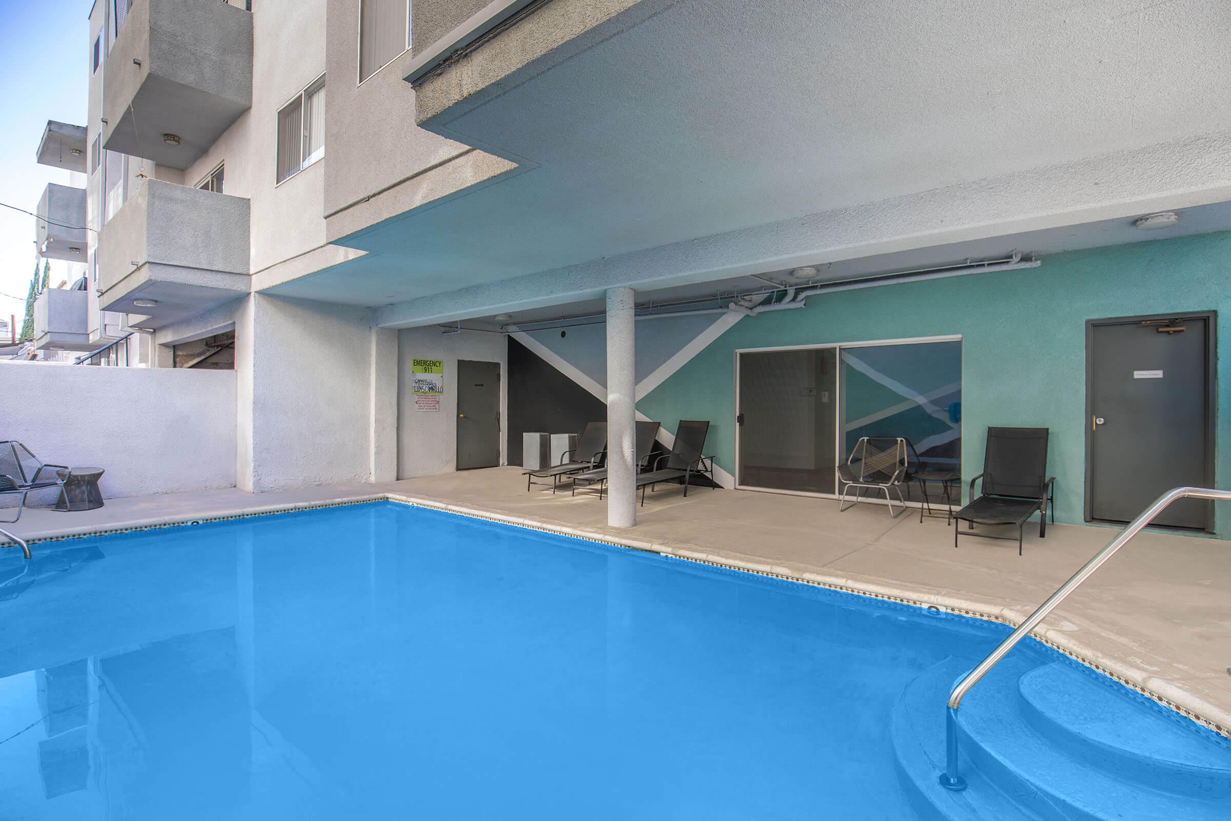 A swimming pool area featuring clear blue water, surrounded by lounge chairs. The pool is located under an overhang of a building, with a wall painted in soft colors. A door leads to a room beside the pool area.