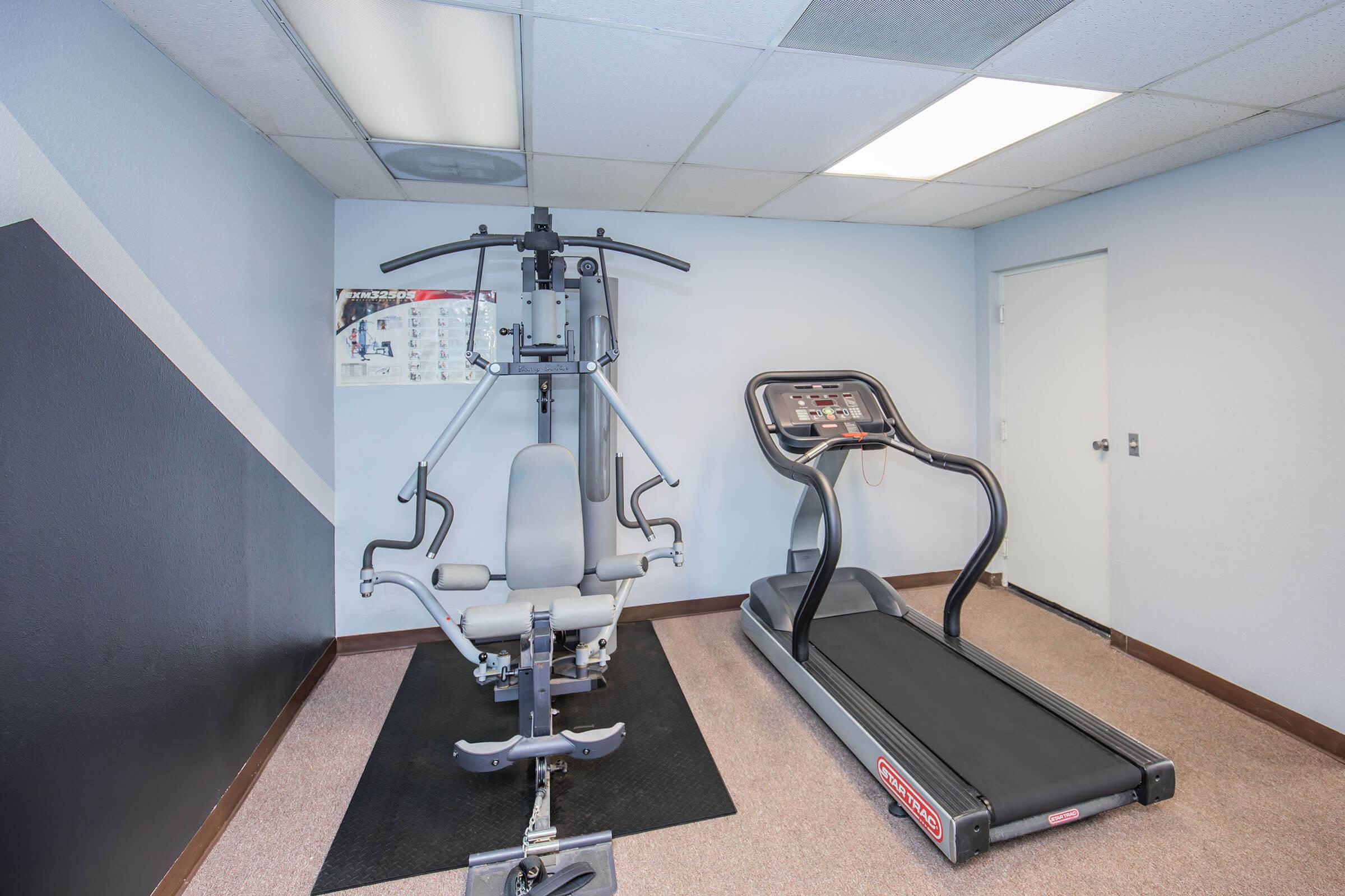 A small home gym setup featuring a weight machine on the left and a treadmill on the right. The room has light-colored walls and a carpeted floor, with a bulletin board picture on the wall in the background. The lighting is bright from ceiling fixtures.