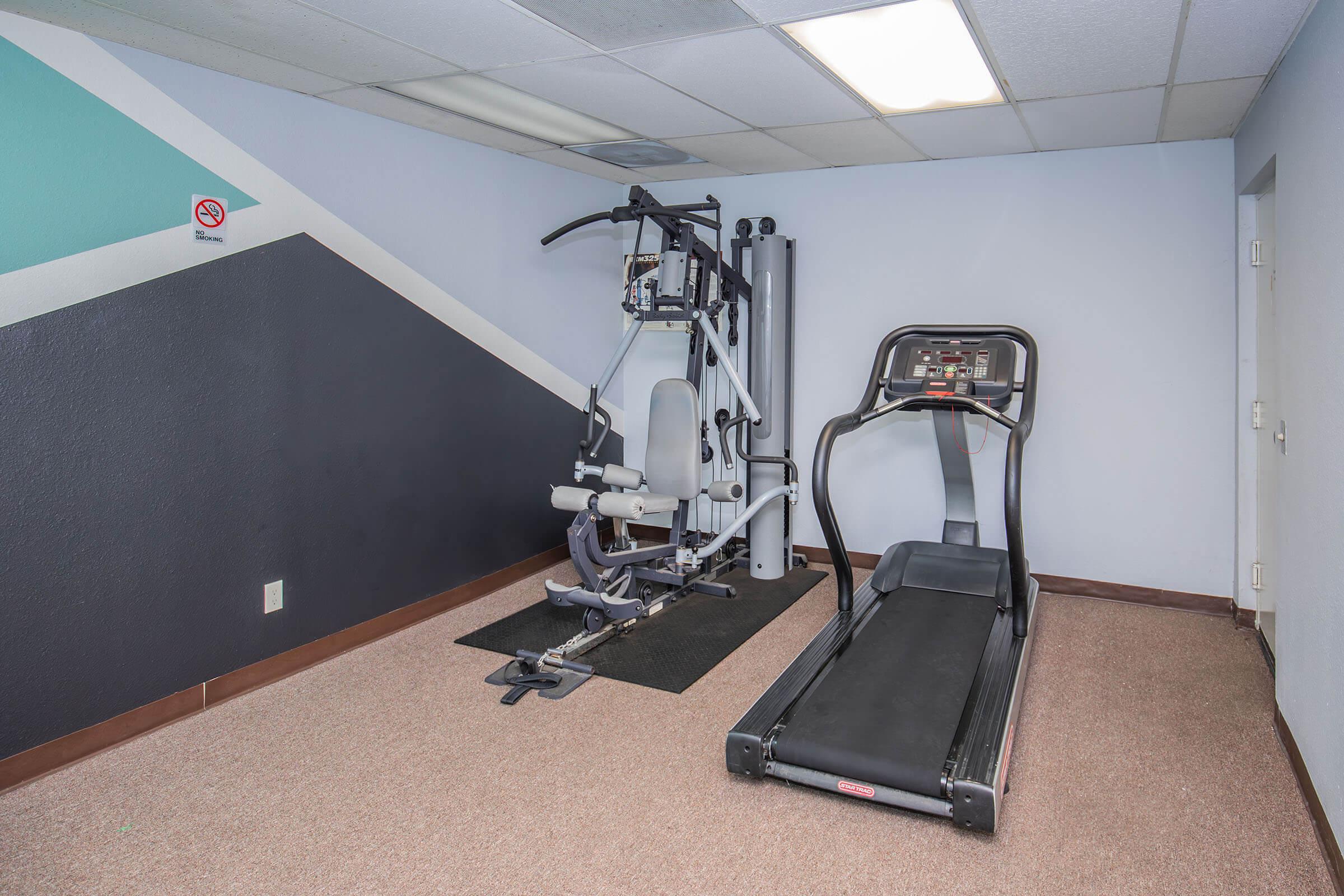 A small fitness room featuring a treadmill and a multi-gym exercise machine. The walls have a geometric design in muted colors, and the floor is carpeted. There are no windows and a no smoking sign is visible on the wall.