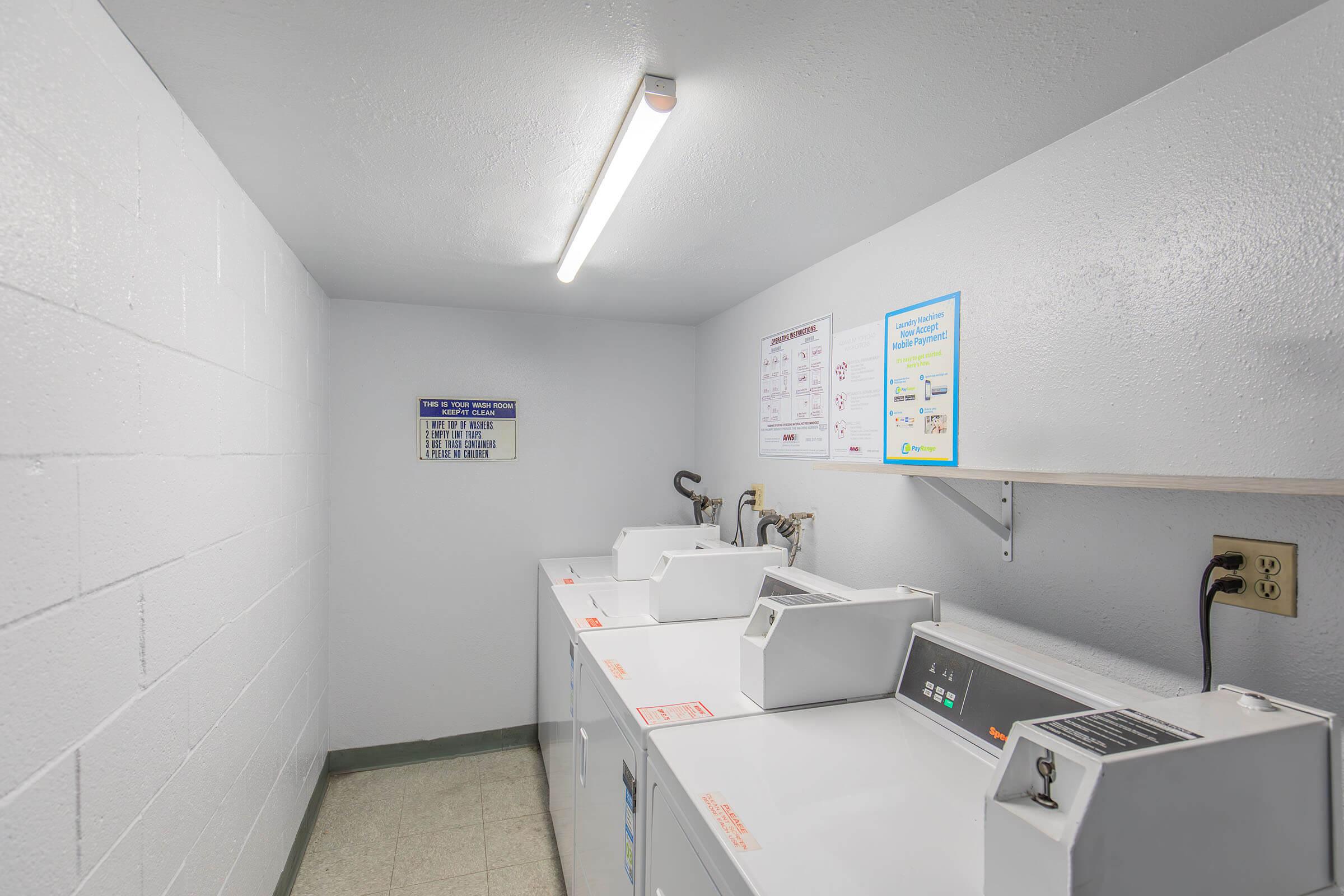 A clean, well-lit laundry room featuring several white washing machines and dryers lined up against a wall. The walls are painted a light gray with white accents, and there are informational signs mounted nearby. A fluorescent light fixture illuminates the space, creating a tidy and functional environment.