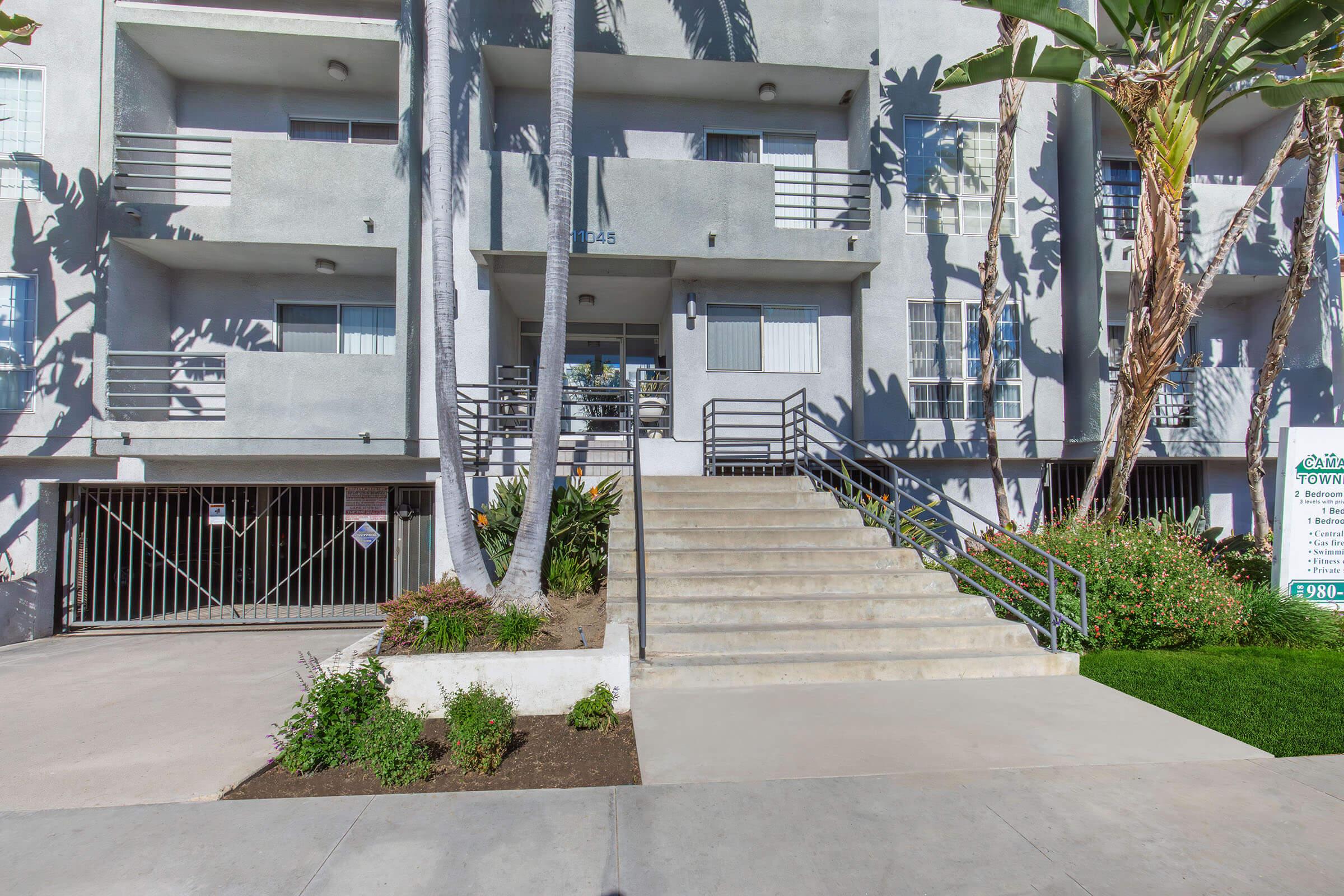 Modern apartment building with a gray exterior. The entrance features steps leading up to sliding glass doors, flanked by palm trees and landscaping. A gated parking area is visible on the left, and a sign stands on the right near a patch of greenery. The setting is bright and well-maintained.