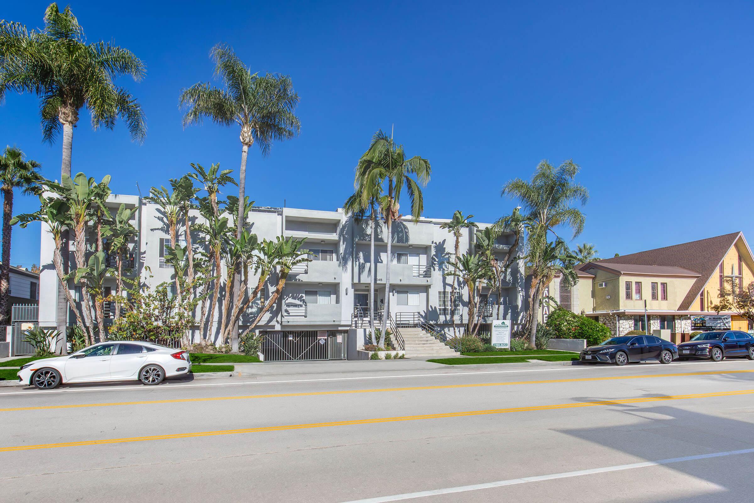 Modern apartment building with palm trees in front, located on a sunny street. The building features a light-colored façade and multiple stories. In the foreground, two cars are parked along the road, with a clear blue sky overhead, creating a vibrant and inviting atmosphere.