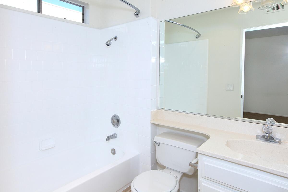 A clean and modern bathroom featuring a white tiled shower-tub combo, a toilet, and a vanity with a sink. There is a large mirror above the sink, reflecting the room's bright and minimalistic design. Natural light enters through a window, enhancing the spacious feel.