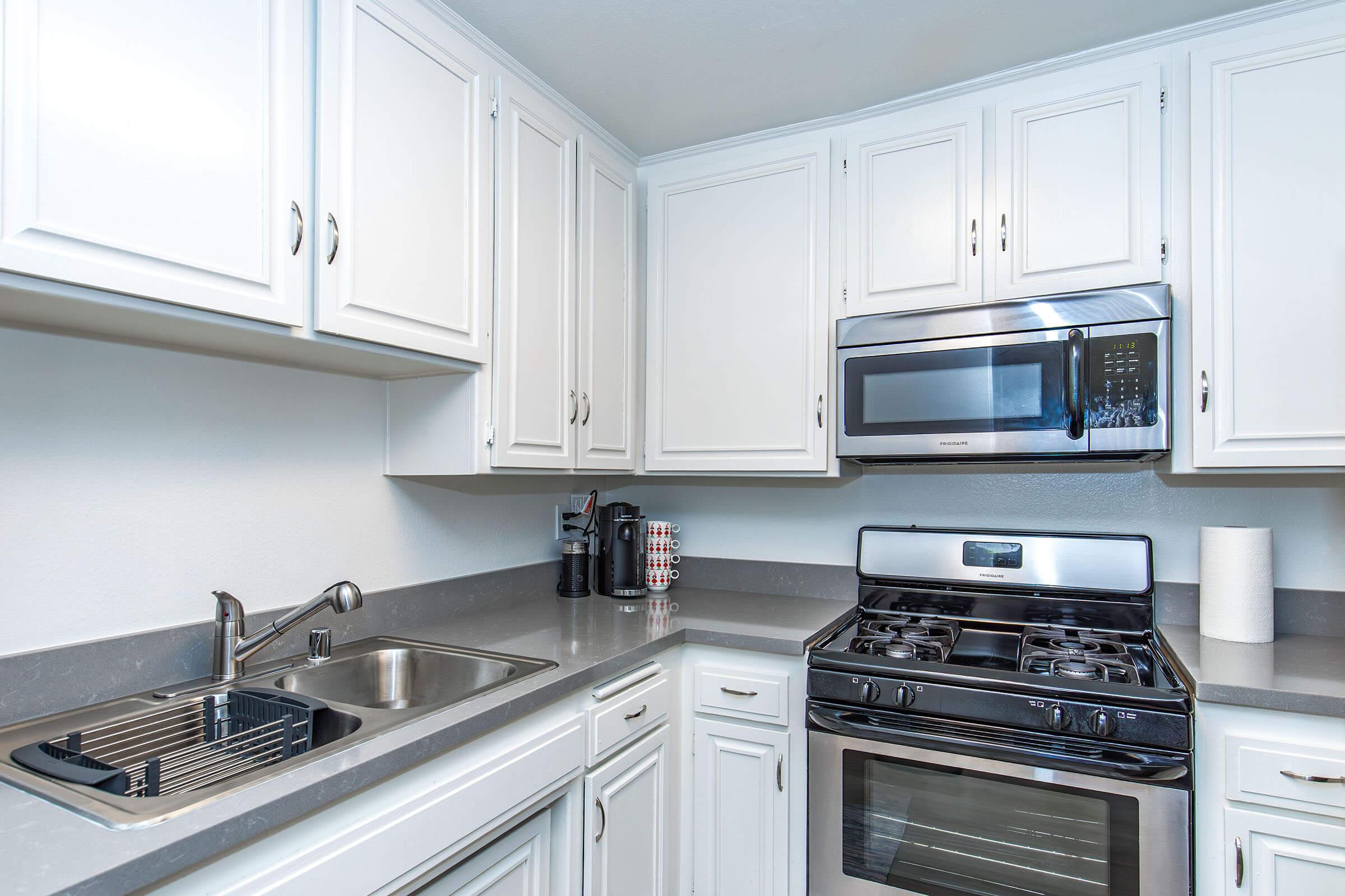Modern kitchen featuring white cabinetry, a gray countertop, a stainless steel sink, a black gas stove, and a microwave. Includes a coffee maker and kitchen accessories, with ample natural light illuminating the space.