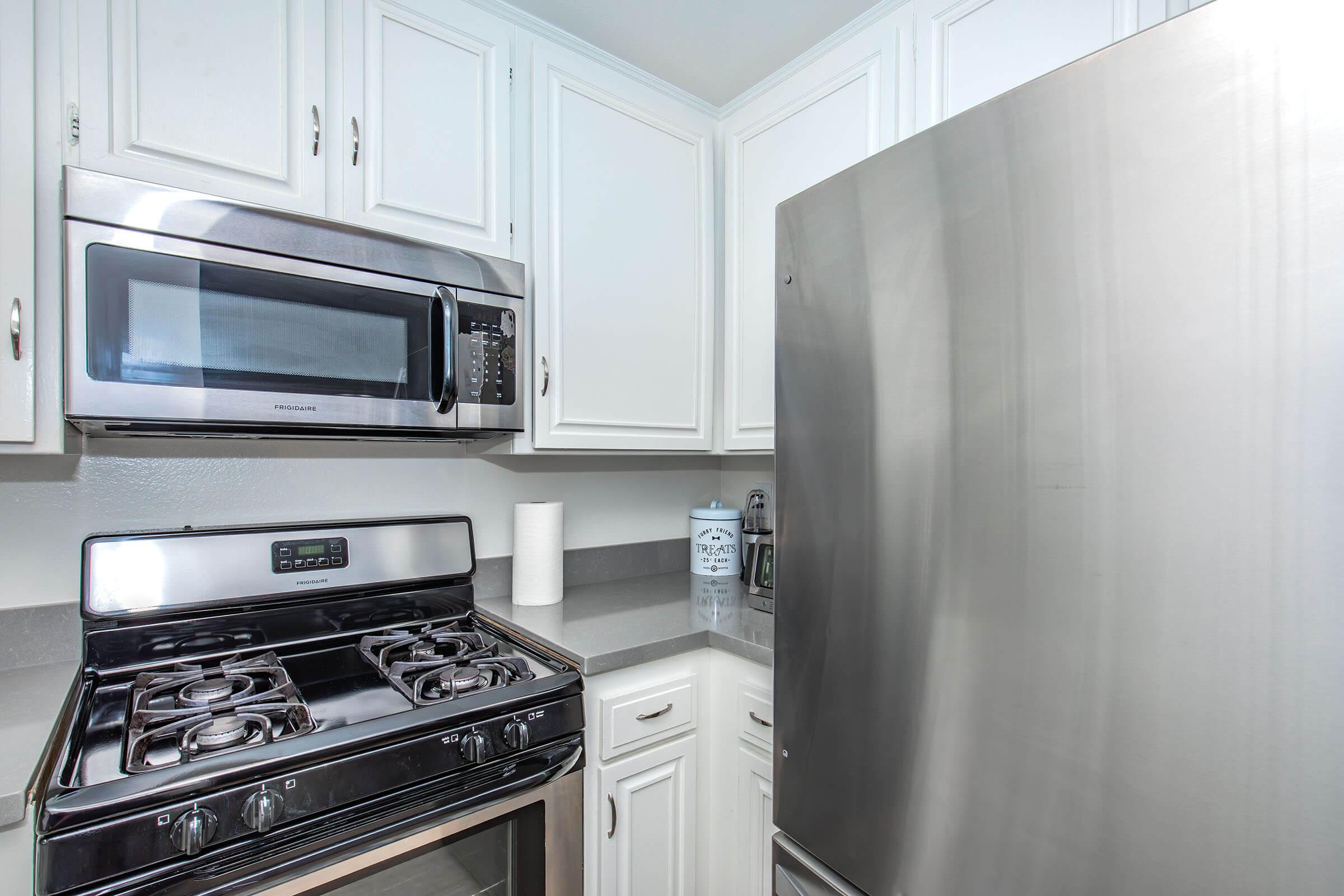 A modern kitchen featuring white cabinetry, a stainless steel refrigerator, a gas stove with an oven, and a microwave above the stove. The countertop is gray, and a roll of paper towels is visible. The overall design is clean and contemporary.