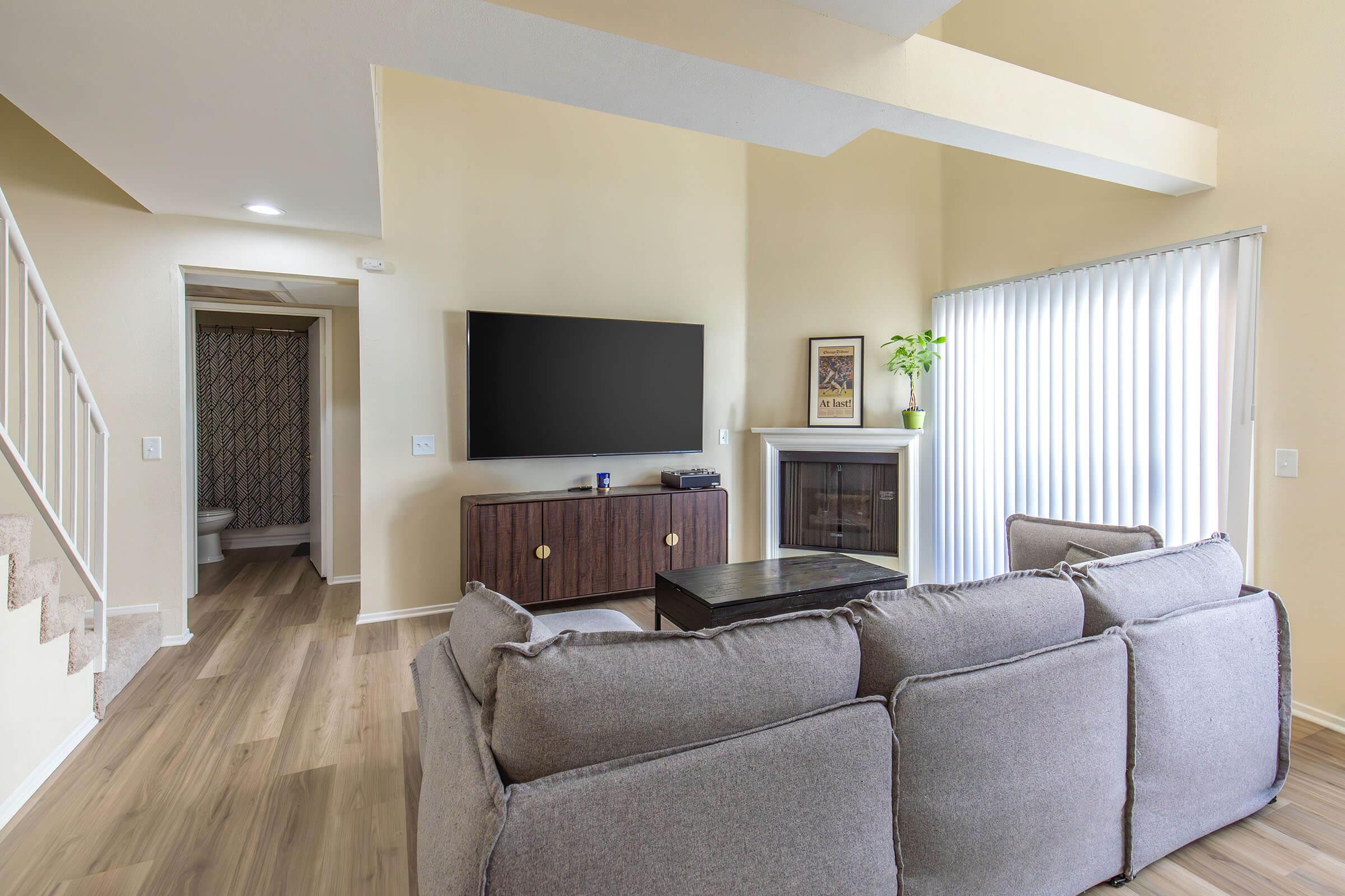 Modern living room featuring a gray sectional sofa facing a large flat-screen TV mounted on a wooden cabinet. Natural light filters through vertical blinds, and a small plant adds a touch of greenery. A staircase can be seen in the background, leading to an upper level, with a visible doorway to a bathroom.