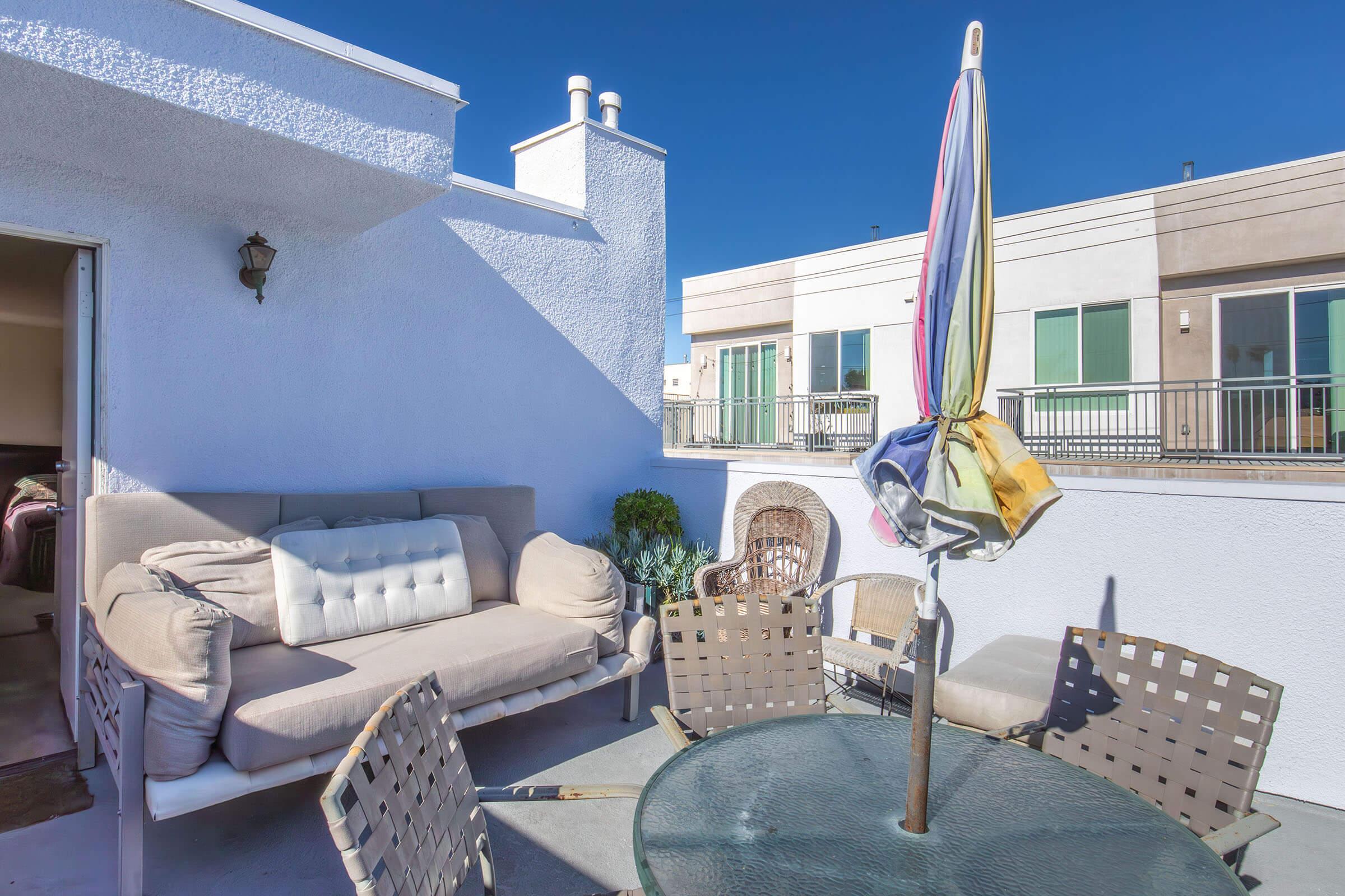 A rooftop terrace with a cream-colored sofa, a round glass table, and several chairs. A colorful umbrella stands in the center of the table. The background features modern buildings and a clear blue sky.