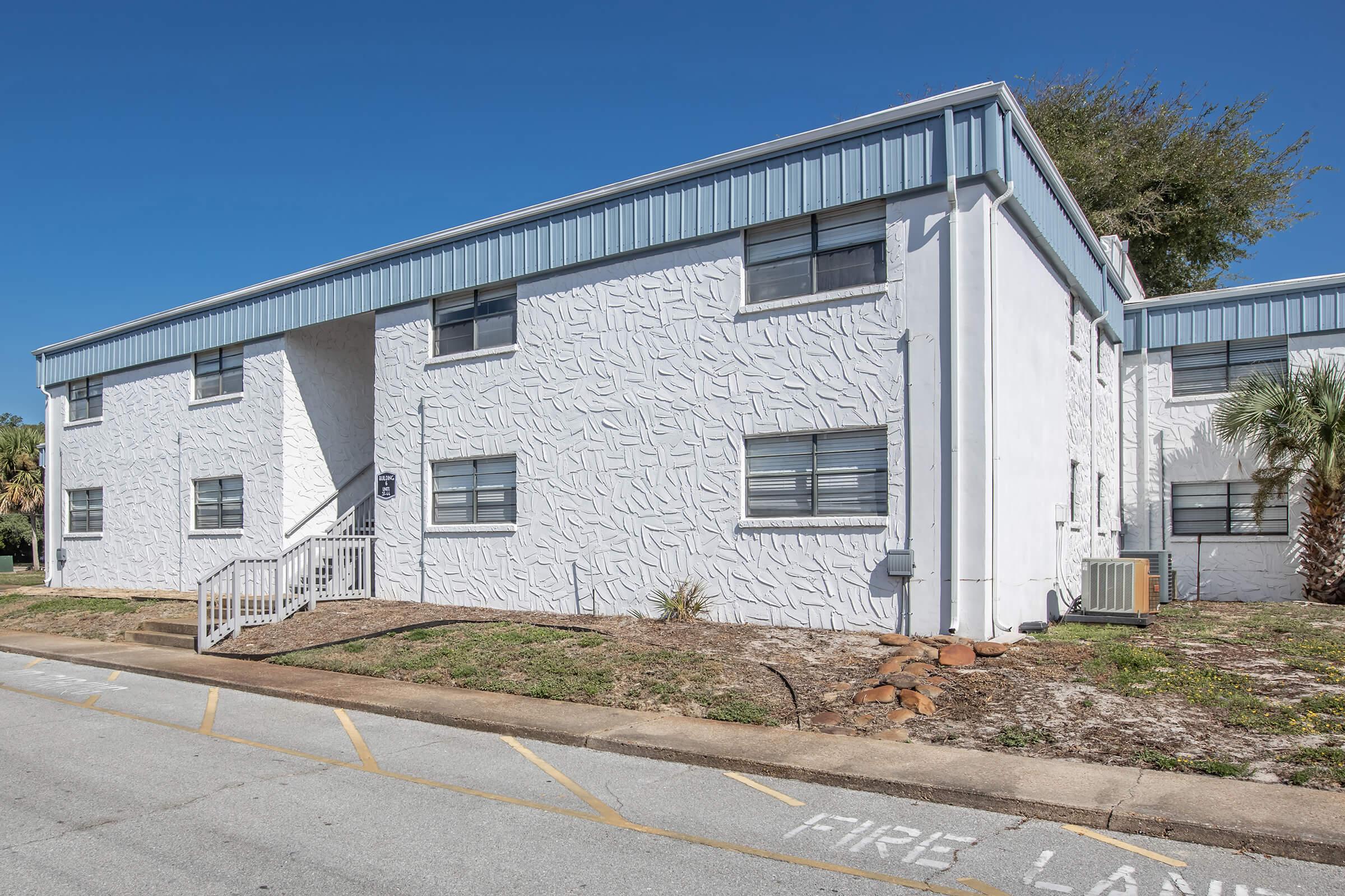 A white multi-unit building with textured exterior, featuring several windows and a small set of stairs leading to the entrance. The surrounding area includes grass and a palm tree, with a clear blue sky overhead. A “FIRE LANE” marking is visible on the pavement in front of the building.