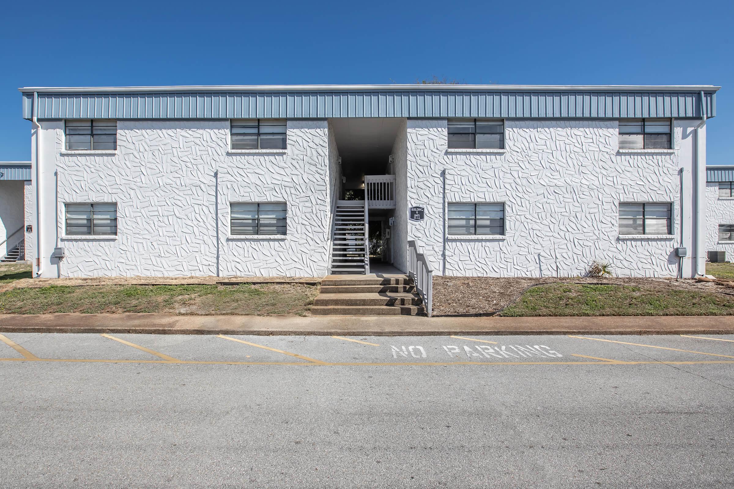 A two-story apartment building with a textured white exterior. The building features several windows and a central staircase leading to the entrance. In front of the building, there are parking spaces with a "No Parking" sign visible on the pavement. Clear blue sky in the background.