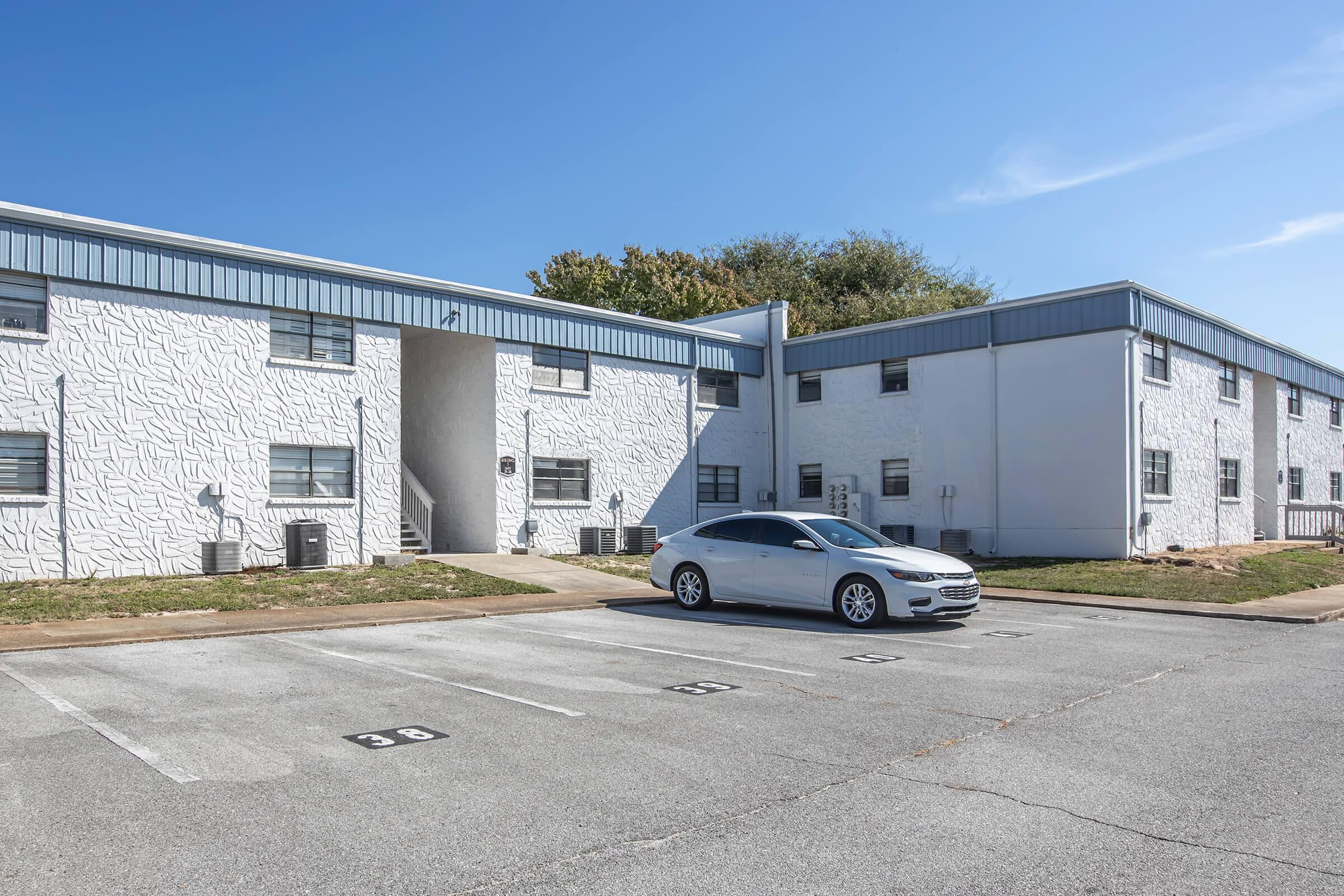 Two white apartment buildings with a textured exterior, one story high, are set in a sunny outdoor environment. A gray sedan is parked in the foreground, with several parking spots visible. The sky is clear with a few scattered clouds, and some greenery is seen in the background.