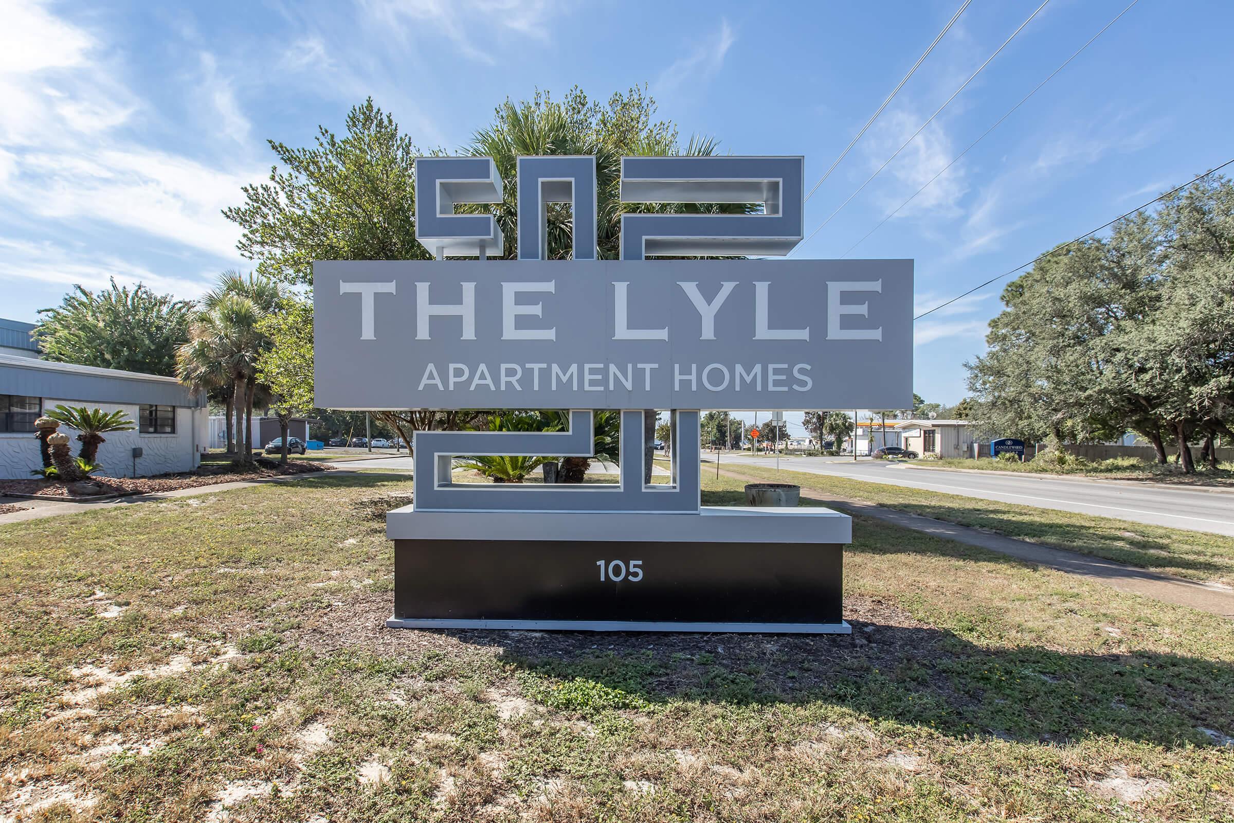 Sign for "The Lyle Apartment Homes" displaying the address 105, surrounded by greenery and palm trees, with a clear blue sky in the background. The sign features a modern design, prominently showcasing the apartment name and address.