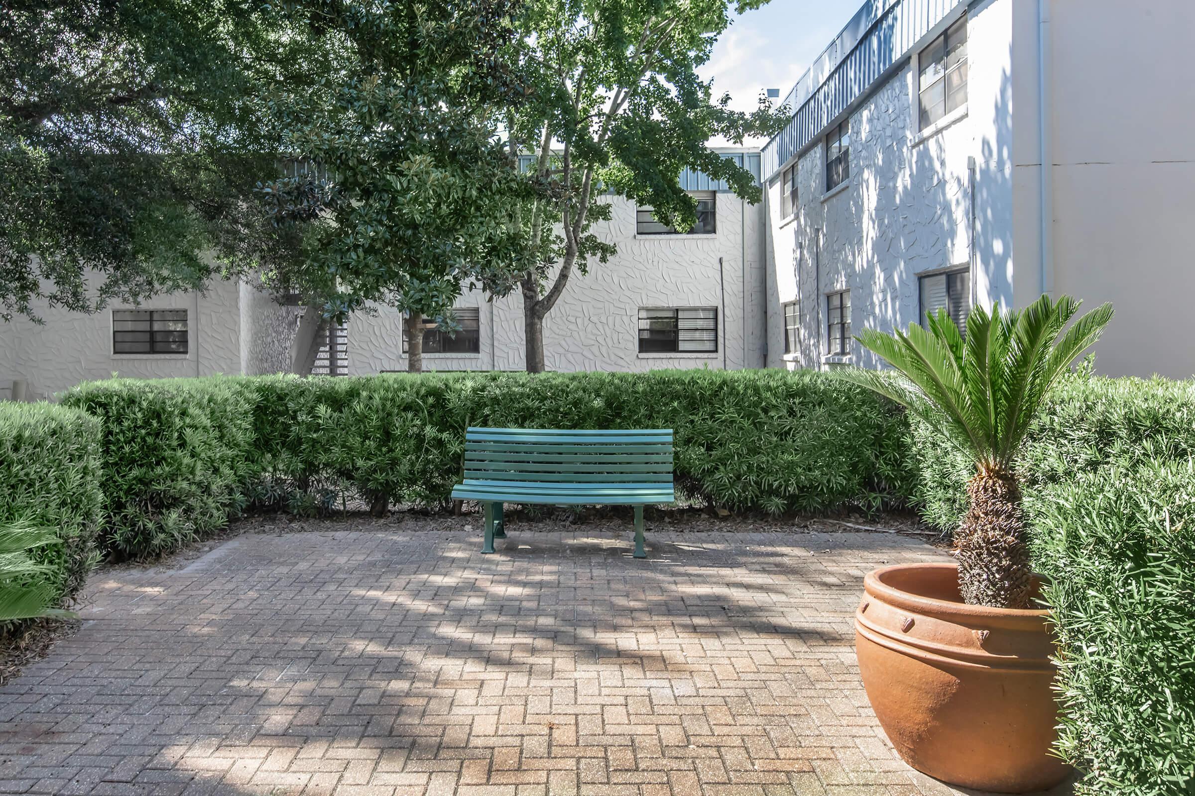 A green park bench surrounded by lush greenery and a large potted plant. In the background, there are white multi-story buildings with windows. The scene is bright and inviting, ideal for relaxation in an outdoor space.