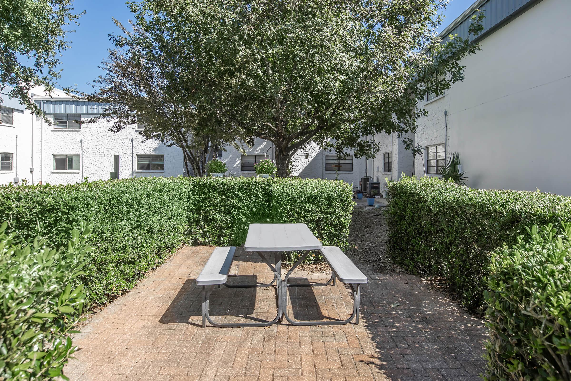 A small outdoor area featuring a grey picnic table with two attached benches, surrounded by neatly trimmed hedges. In the background, there are buildings with a white exterior and a large tree providing shade. The scene is bright and inviting, perfect for casual gatherings.