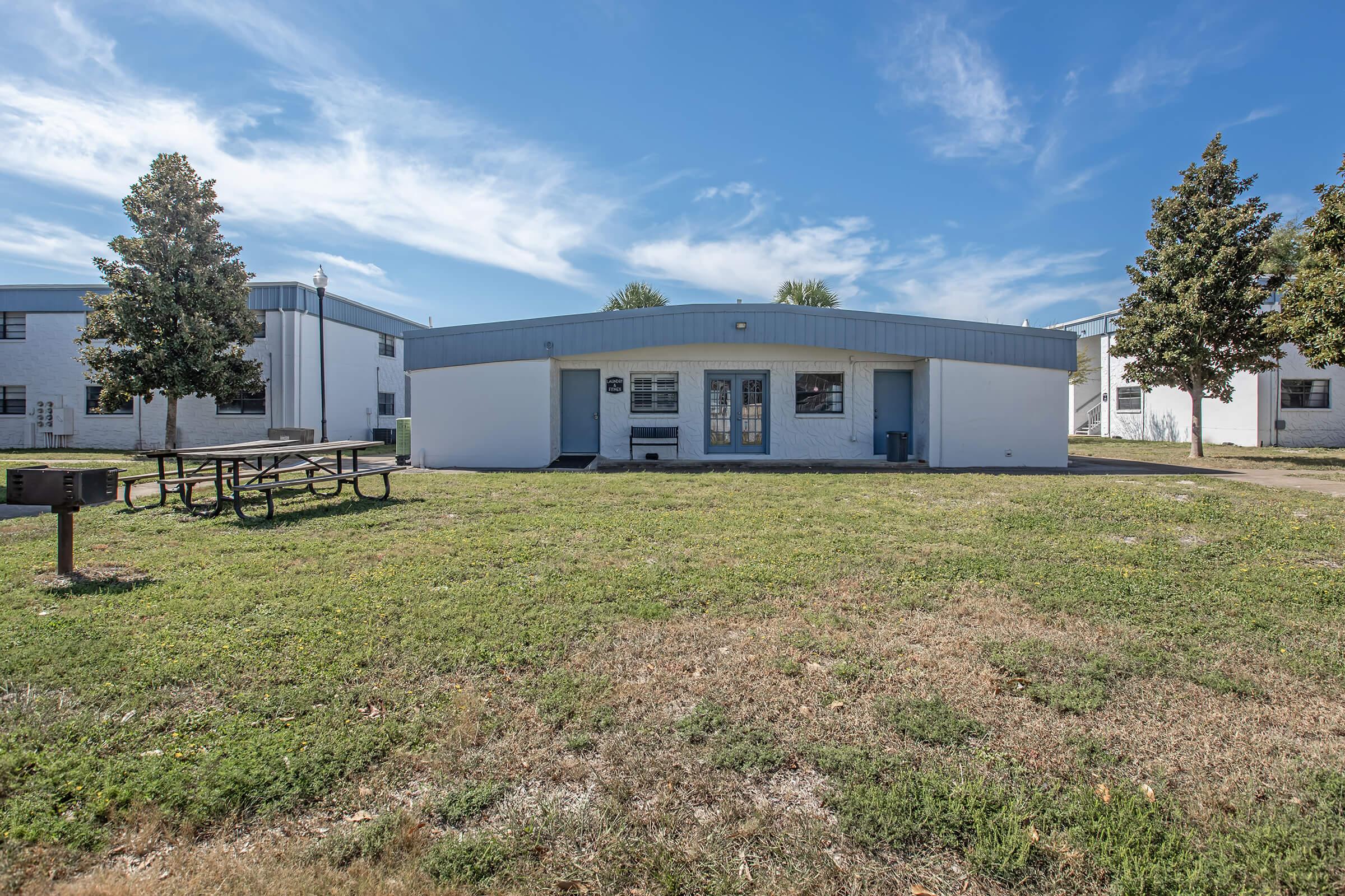 A single-story building with a blue roof, white exterior, and large front windows. In the foreground, a picnic table sits on a grassy area, with a few trees nearby. The sky is bright and partly cloudy, and there are additional buildings visible in the background.