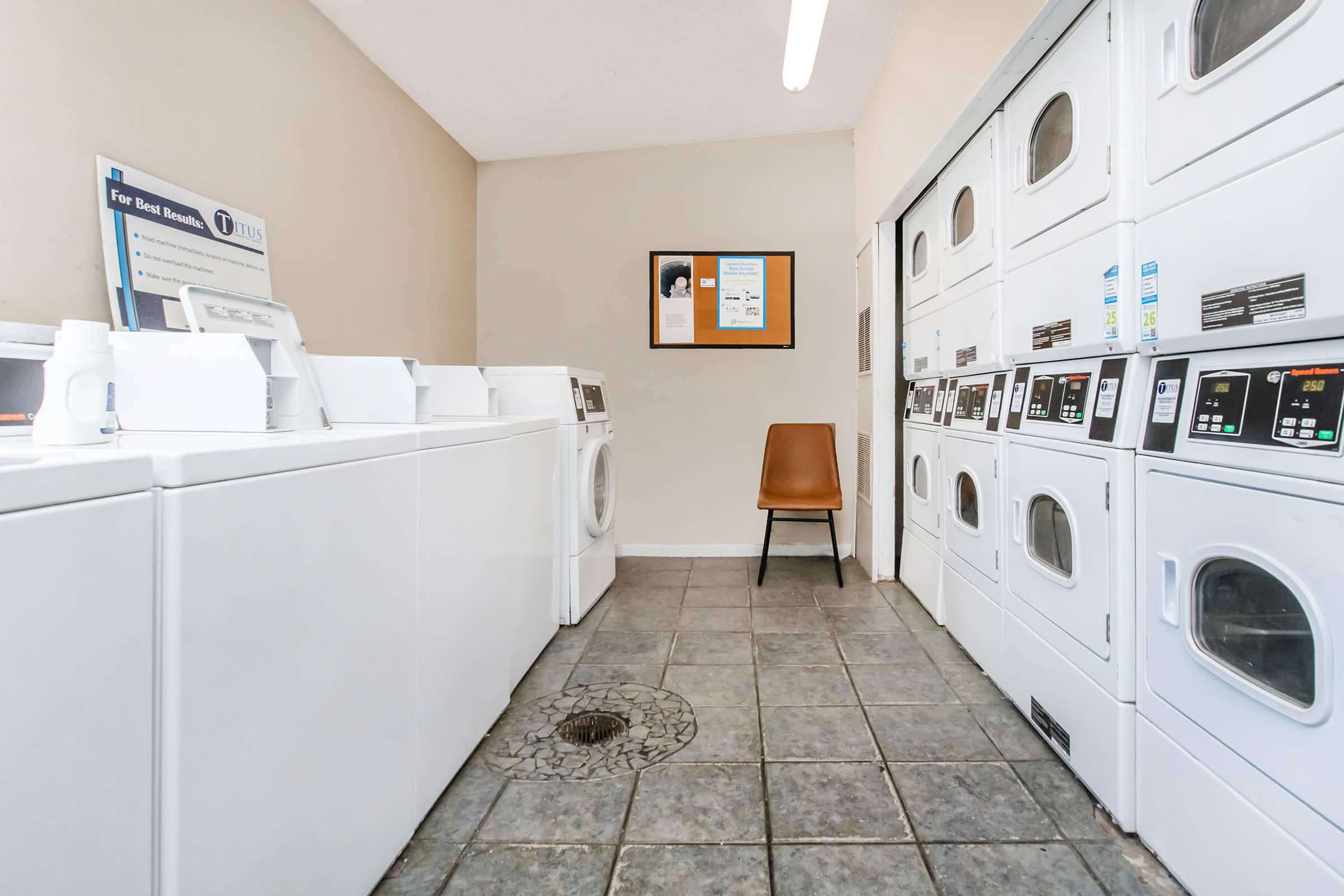 A clean, bright laundry room featuring several white washing machines and dryers neatly aligned. There is a single brown chair against the wall and an informational poster on the wall. The floor is tiled with a circular drain visible in the center. The overall atmosphere is tidy and organized.