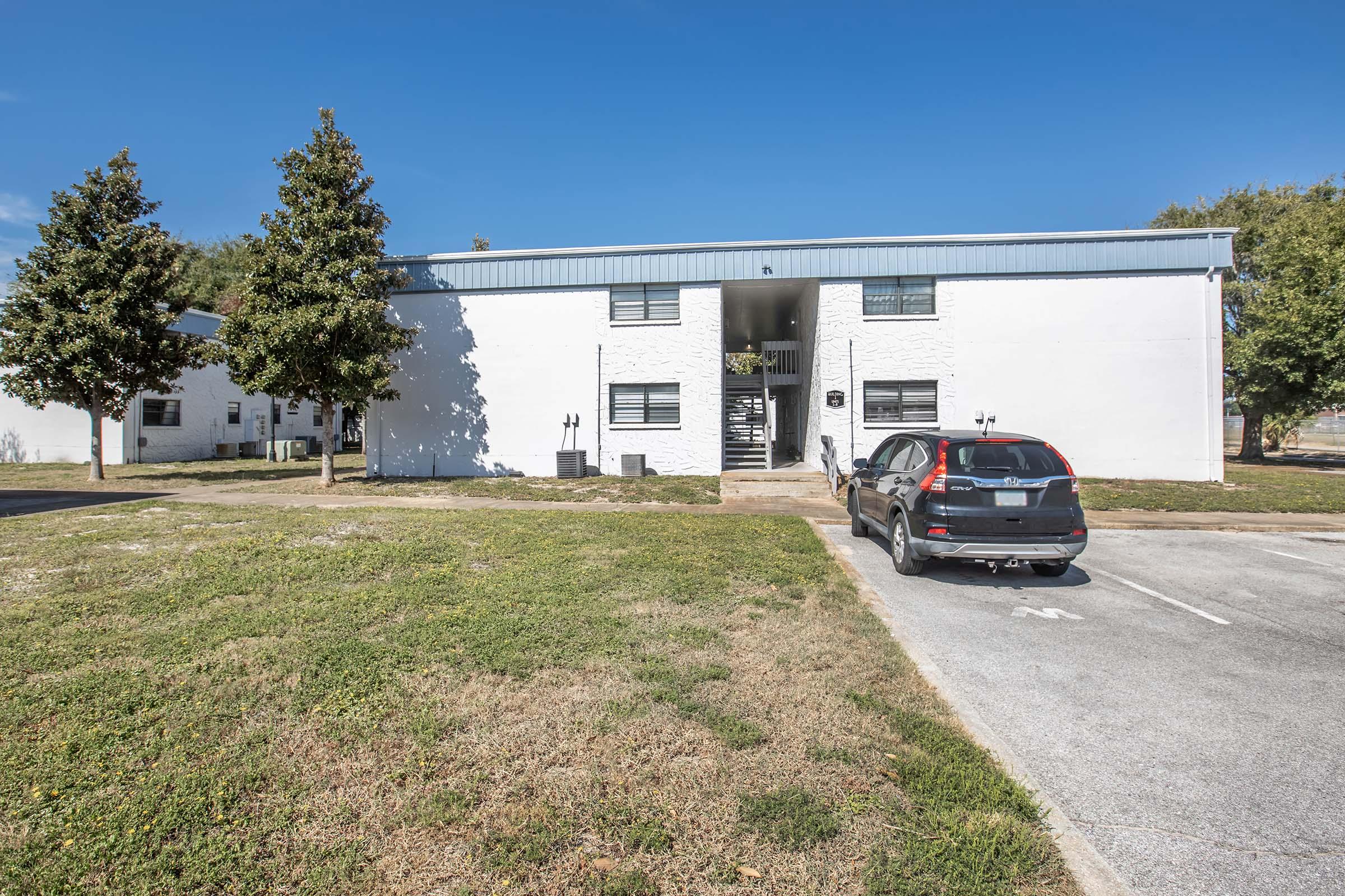A two-story white apartment building with a blue roof, featuring large windows and a staircase on the front. In the foreground, there is a black SUV parked in a designated spot, and grassy areas surround the building under a clear blue sky.
