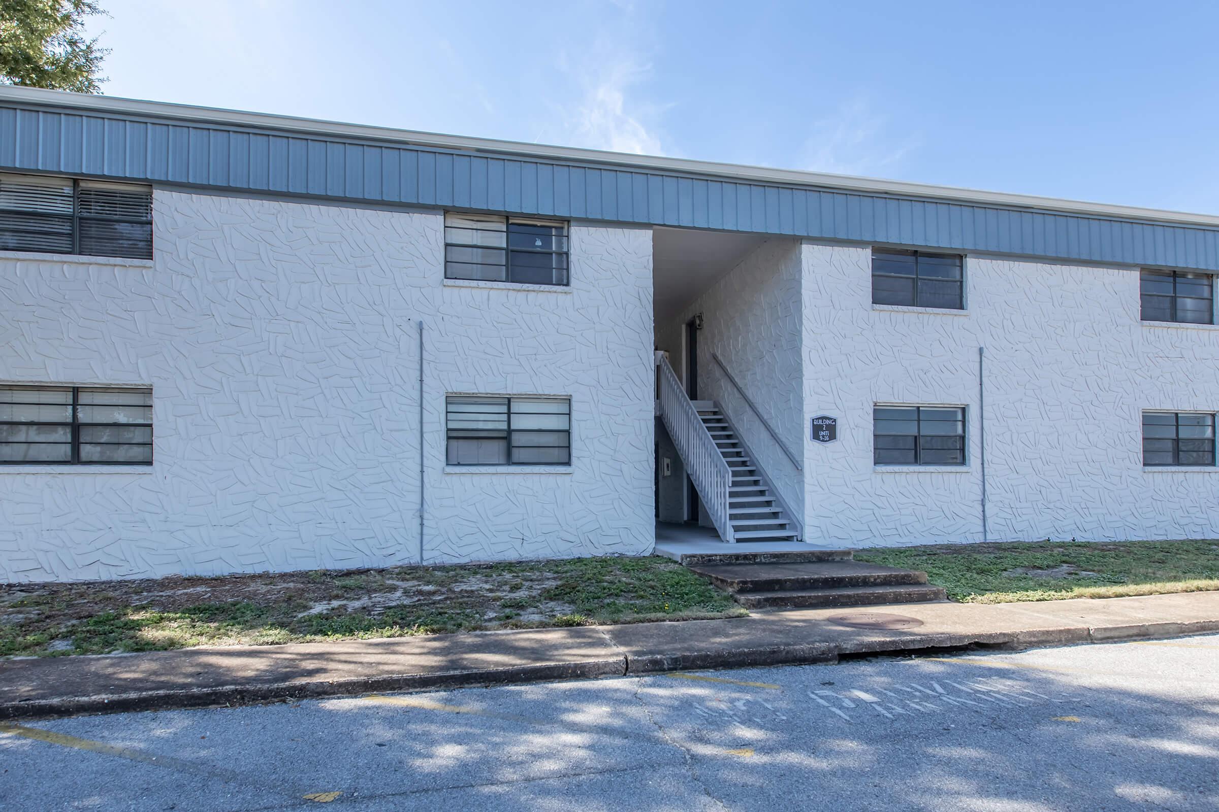 A two-story white apartment building with a textured exterior. The front features multiple windows and a set of stairs leading to the entrance. Surrounding area is a grassy space with a concrete pathway and parking nearby. Clear blue sky in the background.