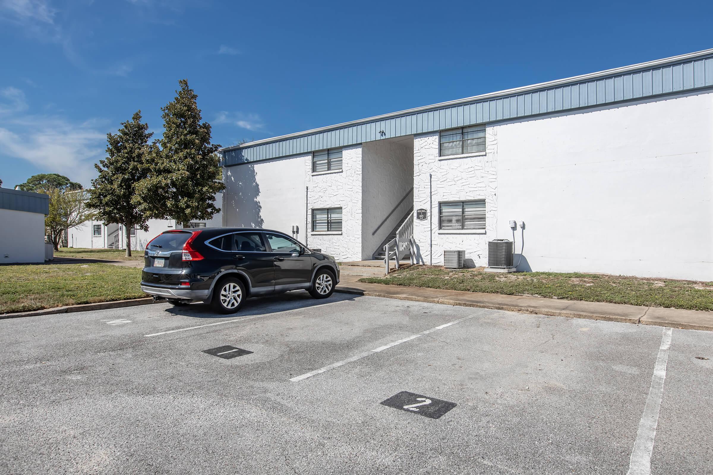 A sunlit view of a parking lot in front of a two-story apartment building. A black SUV is parked in a designated space, while the building features white walls and multiple windows. Grass and a tree are visible in the foreground, with clear blue skies overhead.