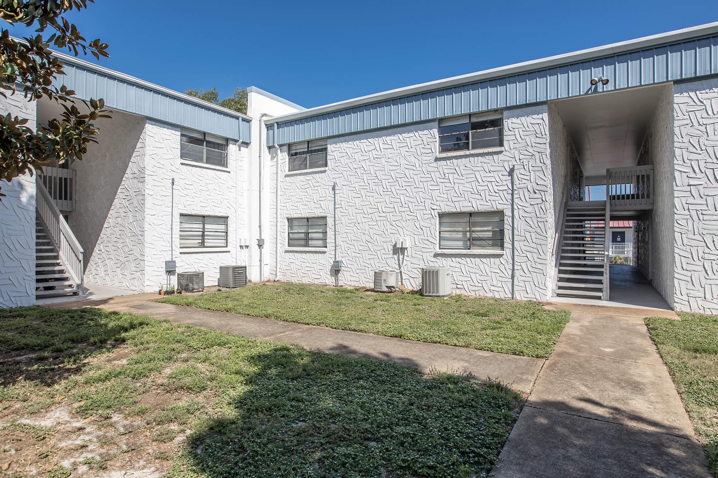Two-story apartment complex featuring white textured walls and blue trim. The courtyard includes a grassy area and concrete pathways leading to open staircases. Air conditioning units are visible on the ground level. It's a clear day with a blue sky.