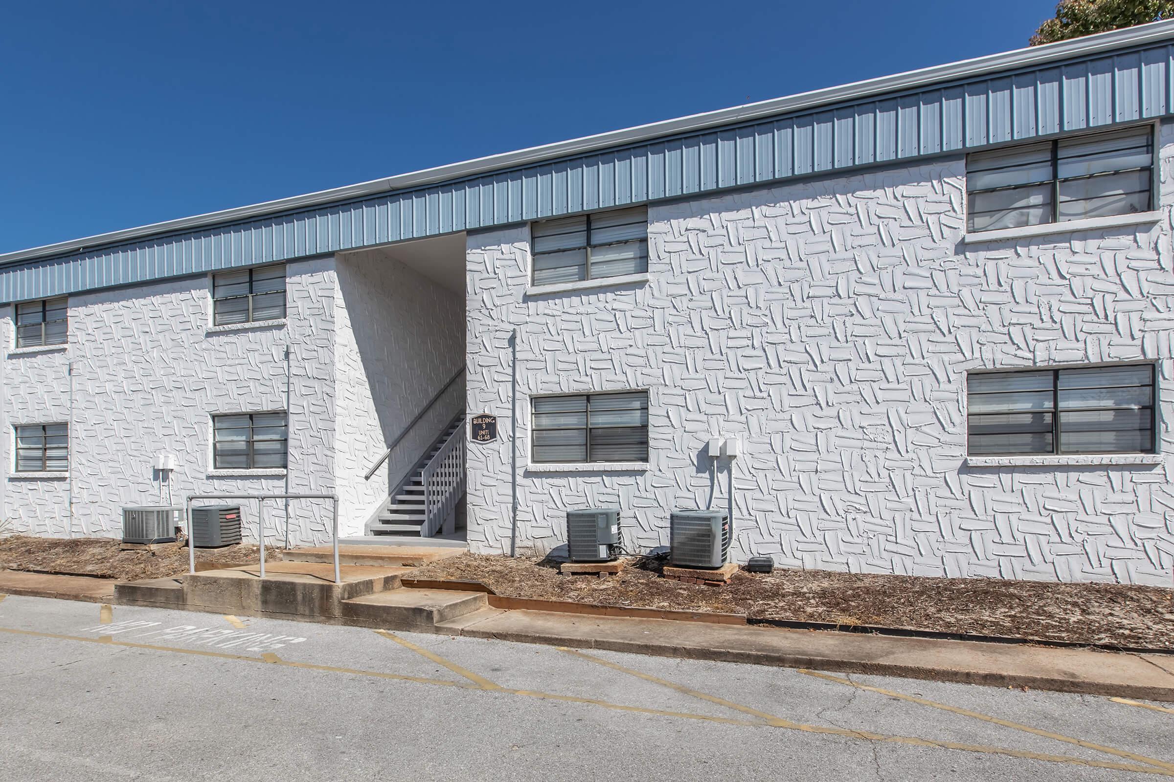 A two-story apartment building with a textured white exterior and blue metal roof. The front features an entrance with stairs leading to the upper floor. Air conditioning units are visible on the ground level, and the surrounding area is clean with gravel and a parking space marked for no parking. Clear blue sky above.