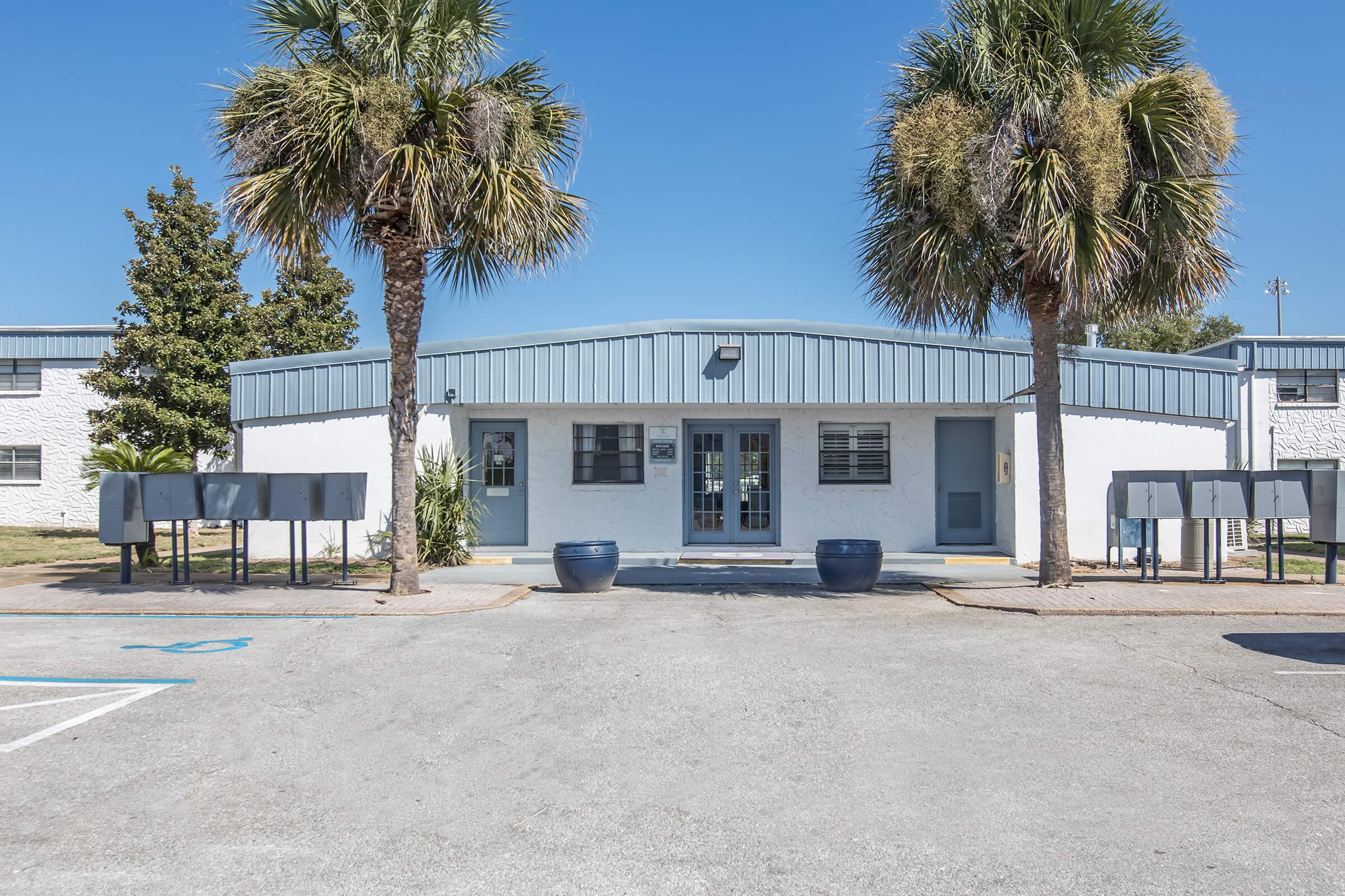 Entrance of a low-rise building with a blue roof and white exterior. Flanked by two palm trees, the front features double doors and several mailboxes lined up. The parking area is visible in the foreground under a clear blue sky.