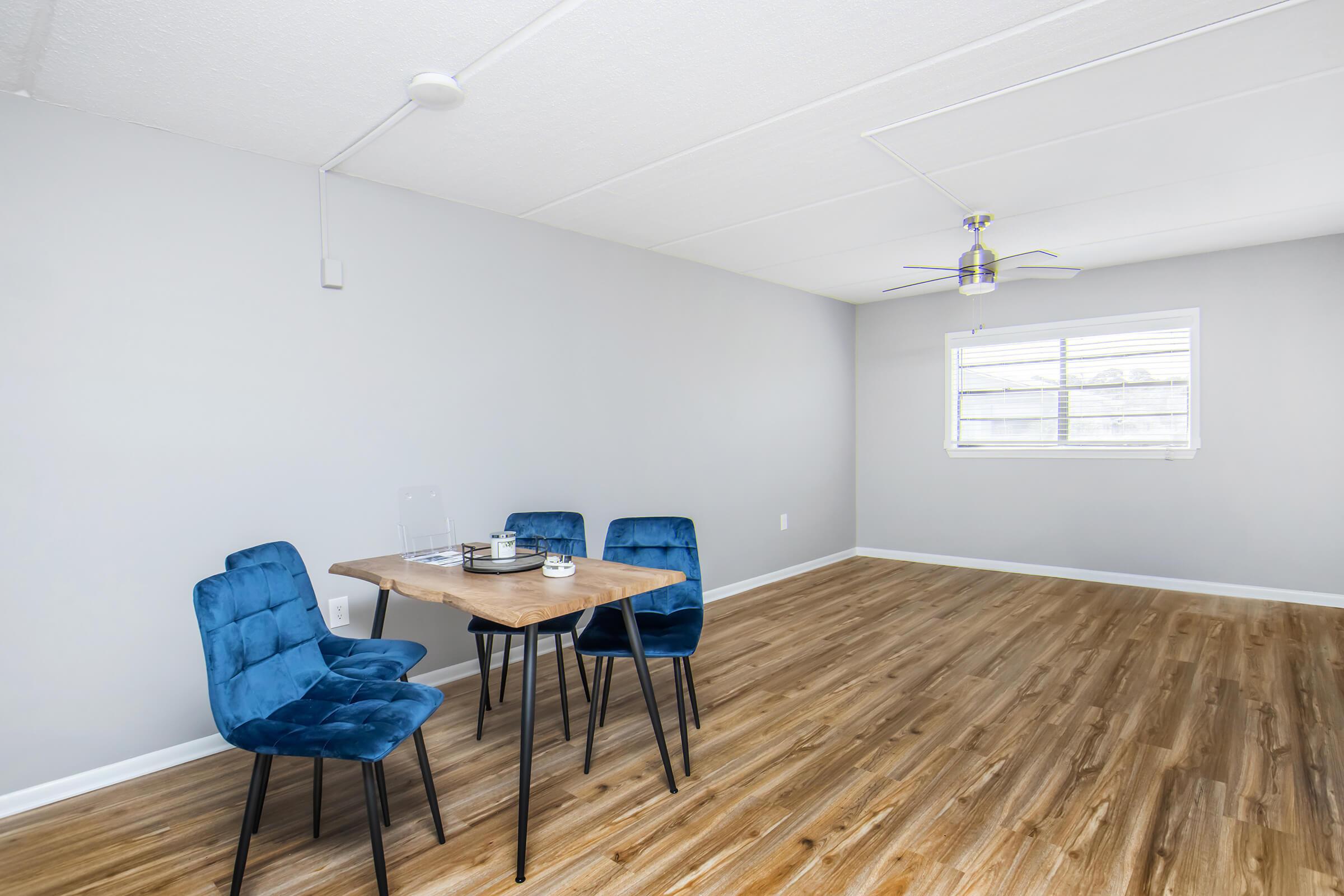 A modern, empty room with light gray walls and wood-like flooring. In the center, there is a small wooden dining table with three blue upholstered chairs around it. A ceiling fan hangs above, and a window provides natural light, enhancing the airy atmosphere.