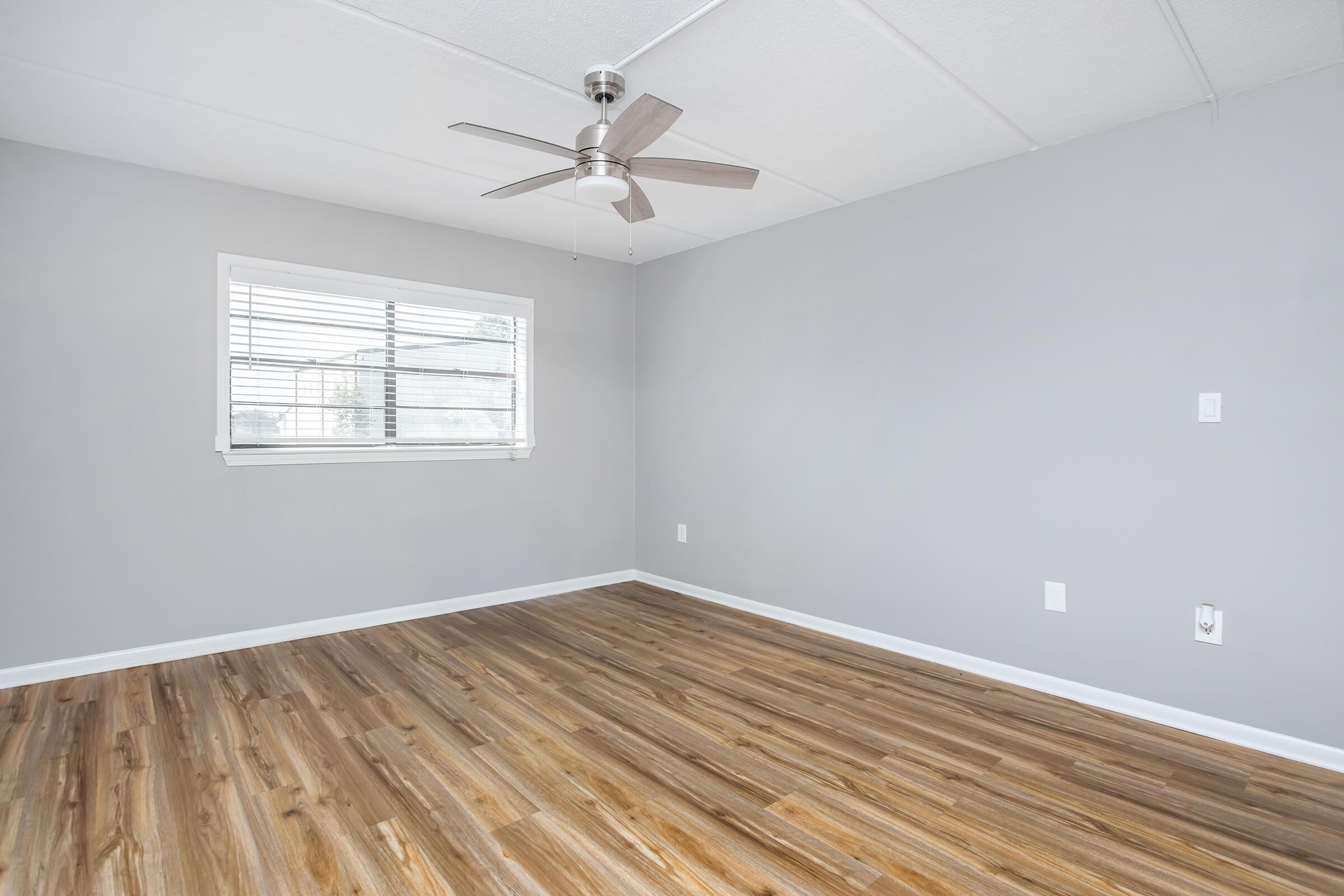 A modern, empty room with grey walls and a wooden floor. There is a ceiling fan in the center and a window with white blinds allowing natural light. The space feels open and bright, ideal for various uses such as a living area or office.