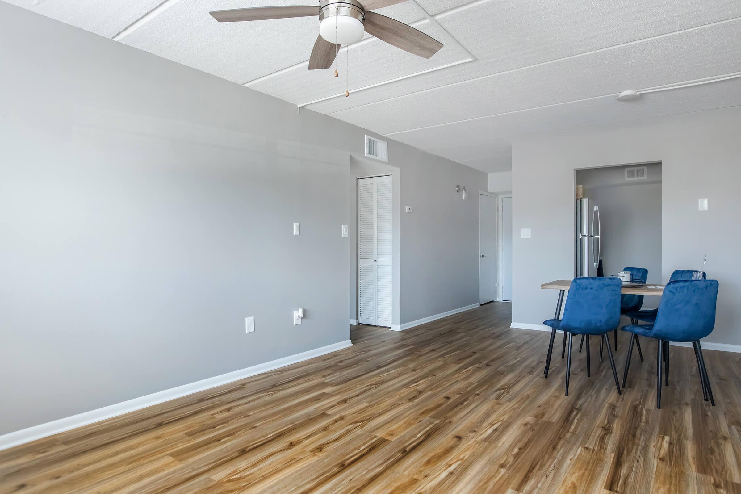 A modern, airy living space featuring light grey walls and wooden flooring. There is a ceiling fan above, and to the right, a dining area with a wooden table and dark blue chairs. A doorway leads to a kitchen with stainless steel appliances visible. The room has a bright and inviting atmosphere.