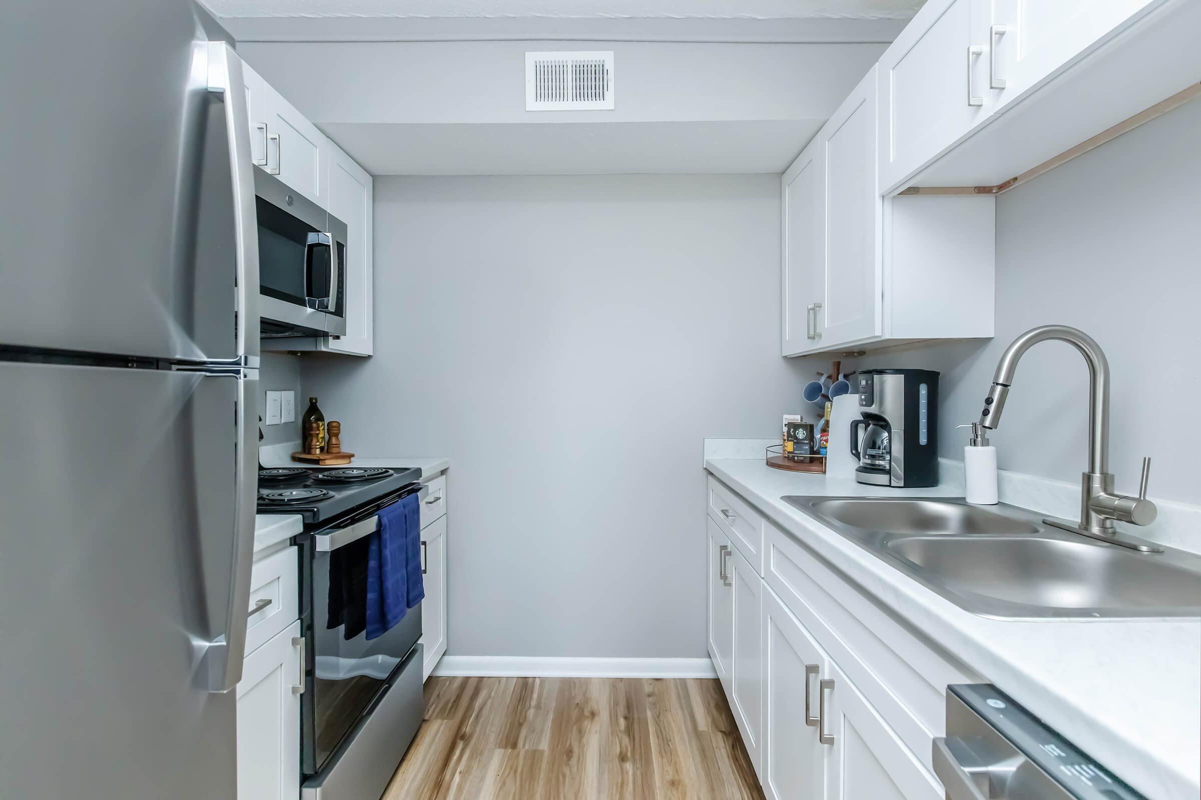 A modern kitchen featuring stainless steel appliances, including a refrigerator and oven. The white cabinets and countertops create a bright, clean look. There's a double sink and a coffee maker on the counter. The flooring is made of light wood, contributing to the contemporary aesthetic of the space.