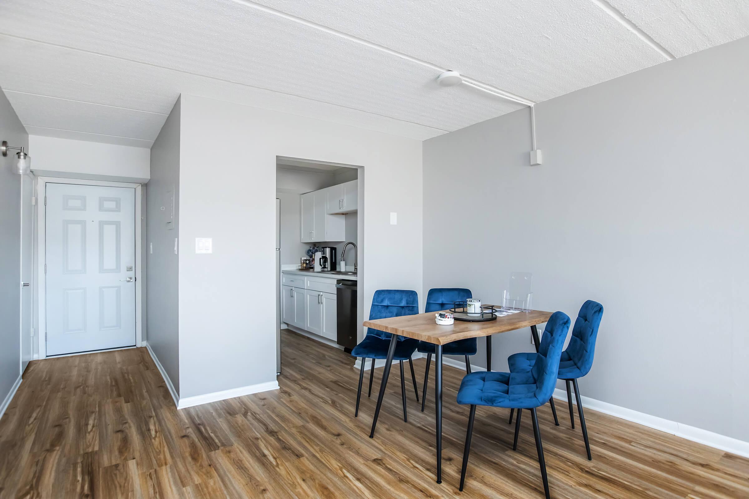 A modern, minimalist dining area featuring a wooden table surrounded by four blue velvet chairs. In the background, a kitchen area is visible through an open doorway. The walls are painted light gray, and the flooring is a light wood. Natural light brightens the space, creating an inviting atmosphere.