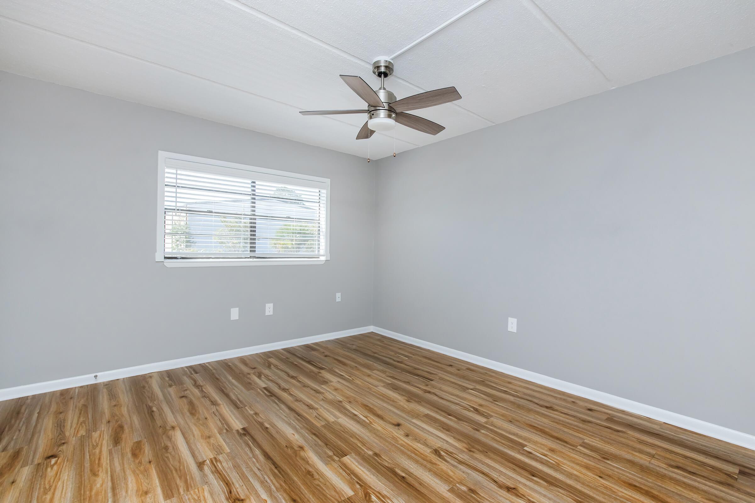 A spacious, empty room featuring gray walls and a wood-like floor. It has a ceiling fan and a window with blinds that allows natural light to enter, enhancing the bright and airy feel of the space. The room is ready for furniture and personal touches.