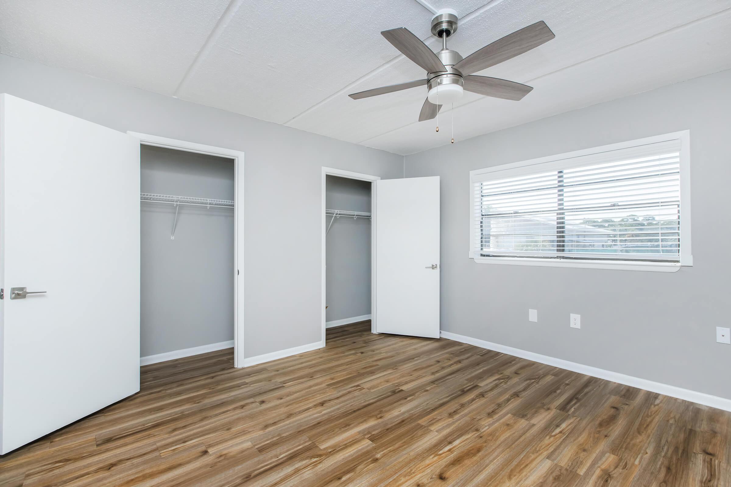 Interior of a modern bedroom featuring light gray walls, a ceiling fan with wooden blades, and two open doors revealing closets. The floor is covered with light wooden laminate. A window with white blinds allows natural light to enter the room.
