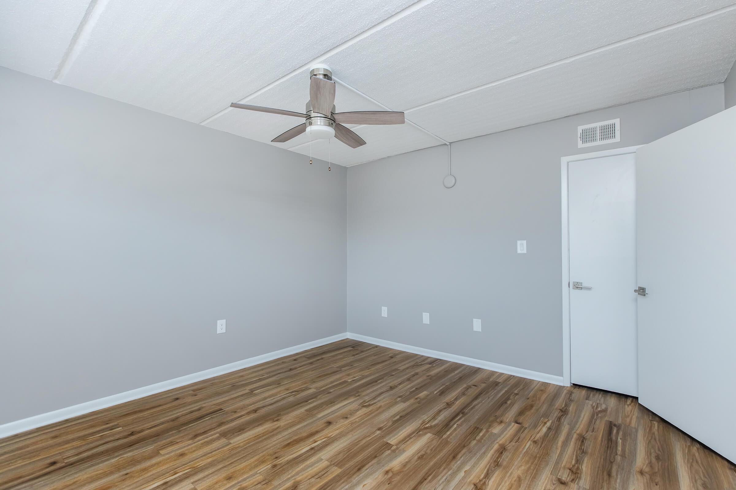 A spacious room featuring light gray walls and a ceiling fan. The floor is finished with wood-like laminate. A white door is visible on the right, and there are several electrical outlets along the walls. The ceiling has a textured finish. Natural light filters in from an unseen window.