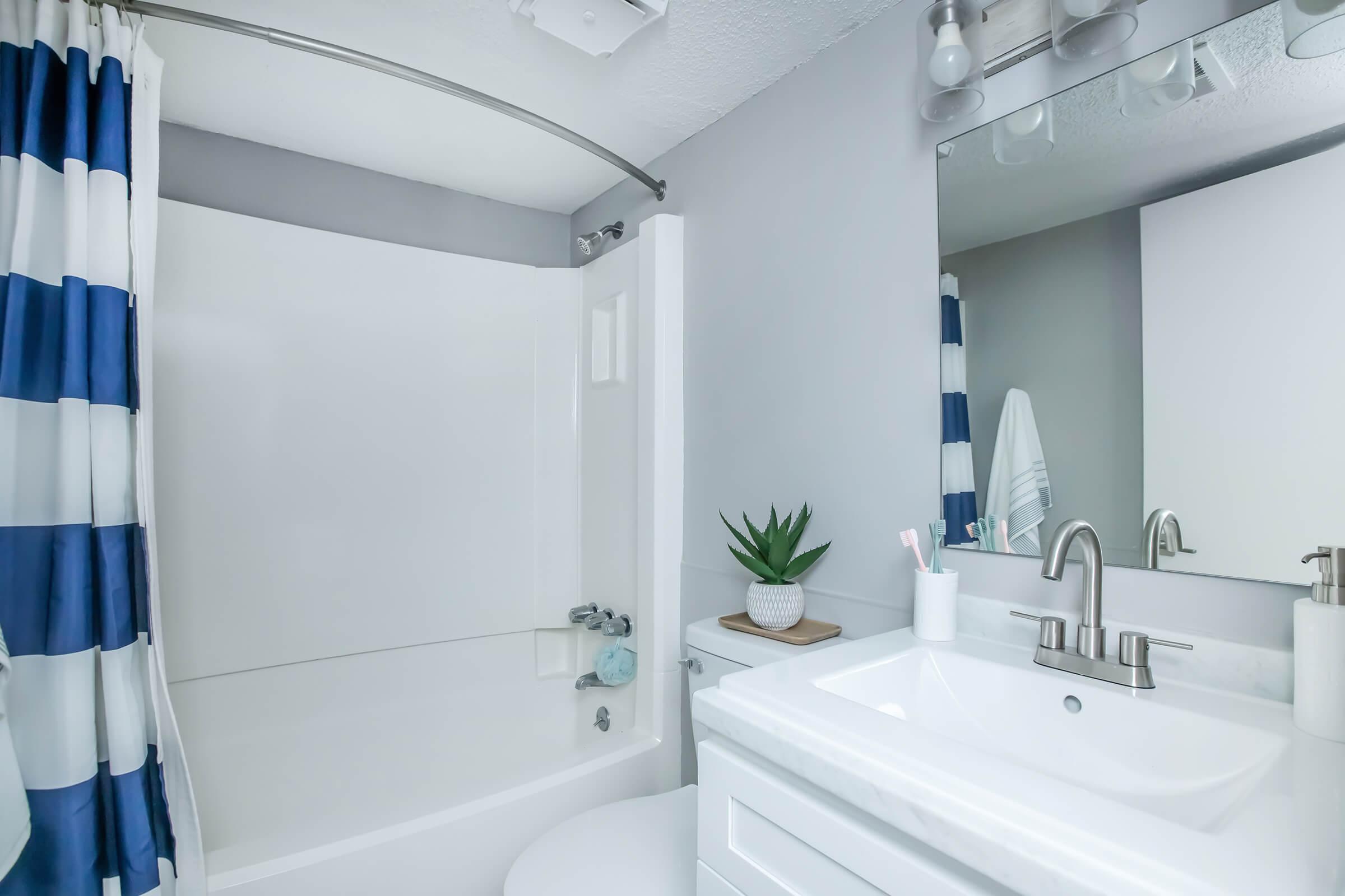A clean and modern bathroom featuring a white bathtub with a blue and white striped shower curtain. A minimalist white sink with a sleek faucet is visible, along with a small potted plant on the countertop. Bright lighting enhances the airy and fresh atmosphere.