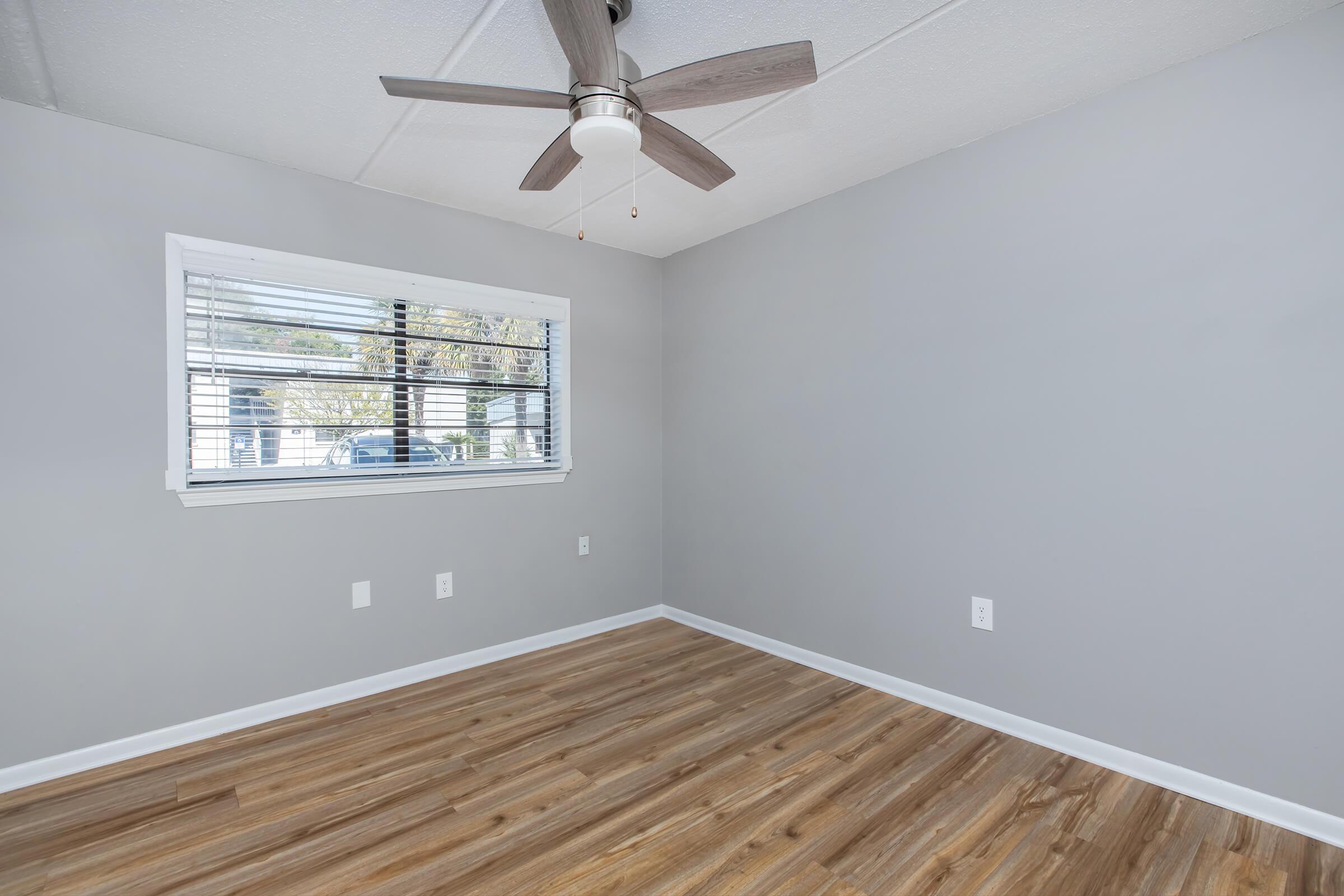 A vacant room featuring light gray walls and a wooden floor. A ceiling fan with wooden blades hangs above, and there is a window with partially closed blinds allowing natural light to enter. The space is unfurnished, creating a clean and simple aesthetic.