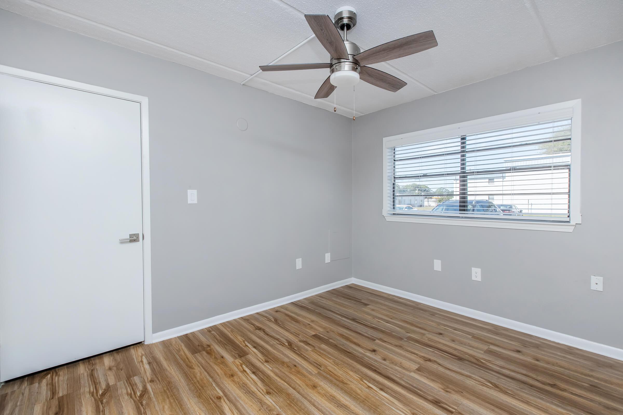 A vacant room featuring light gray walls, a ceiling fan with wooden blades, and tan hardwood flooring. There’s a closed white door to the left and a window with blinds allowing natural light to enter, showcasing a view of the outside. The overall ambiance is bright and minimalistic.