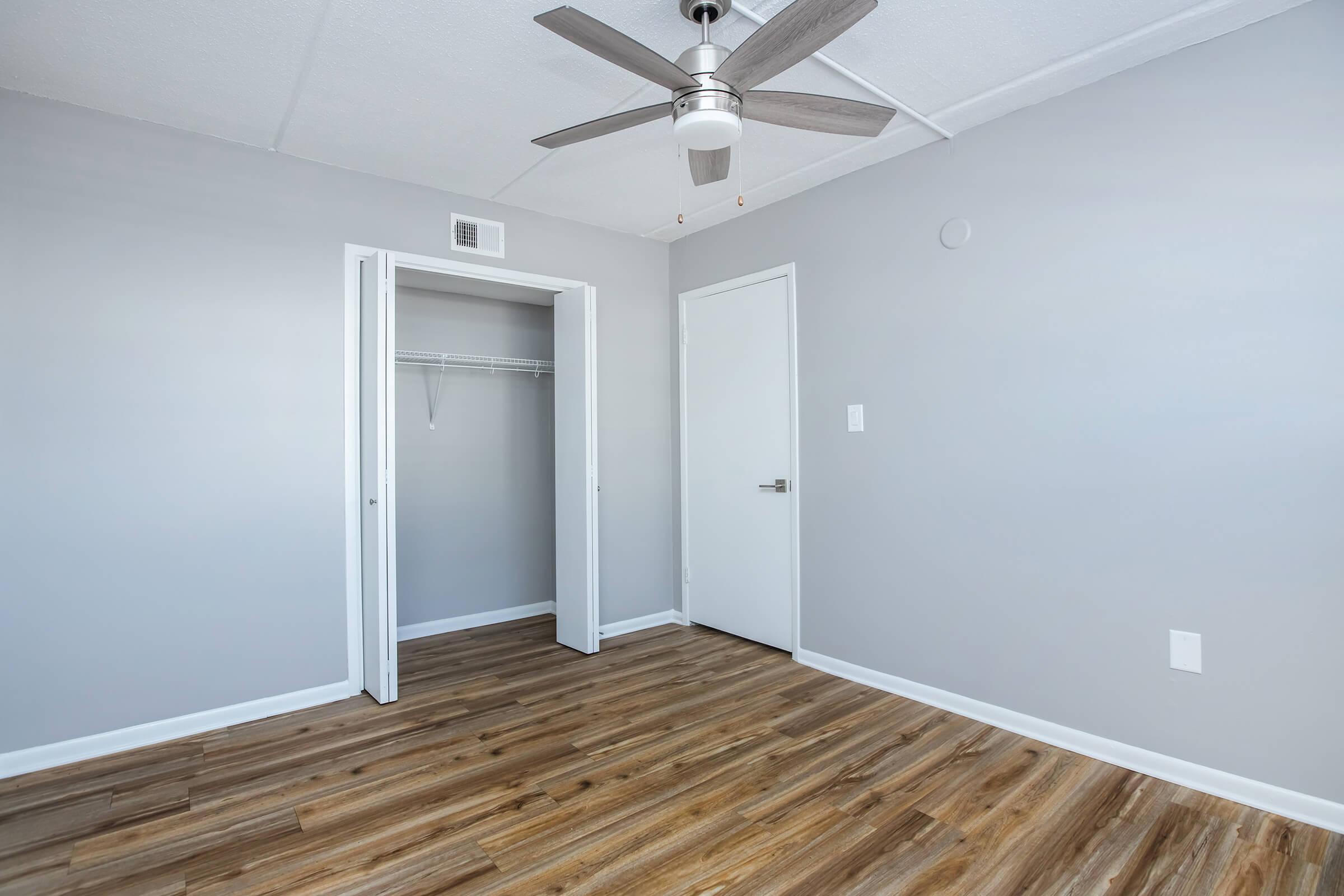 A brightly lit empty room with light gray walls and wooden flooring. There is a ceiling fan with a light fixture and a closet with sliding doors. A closed white door leads to another space. The overall atmosphere is clean and modern, suitable for various interior designs.
