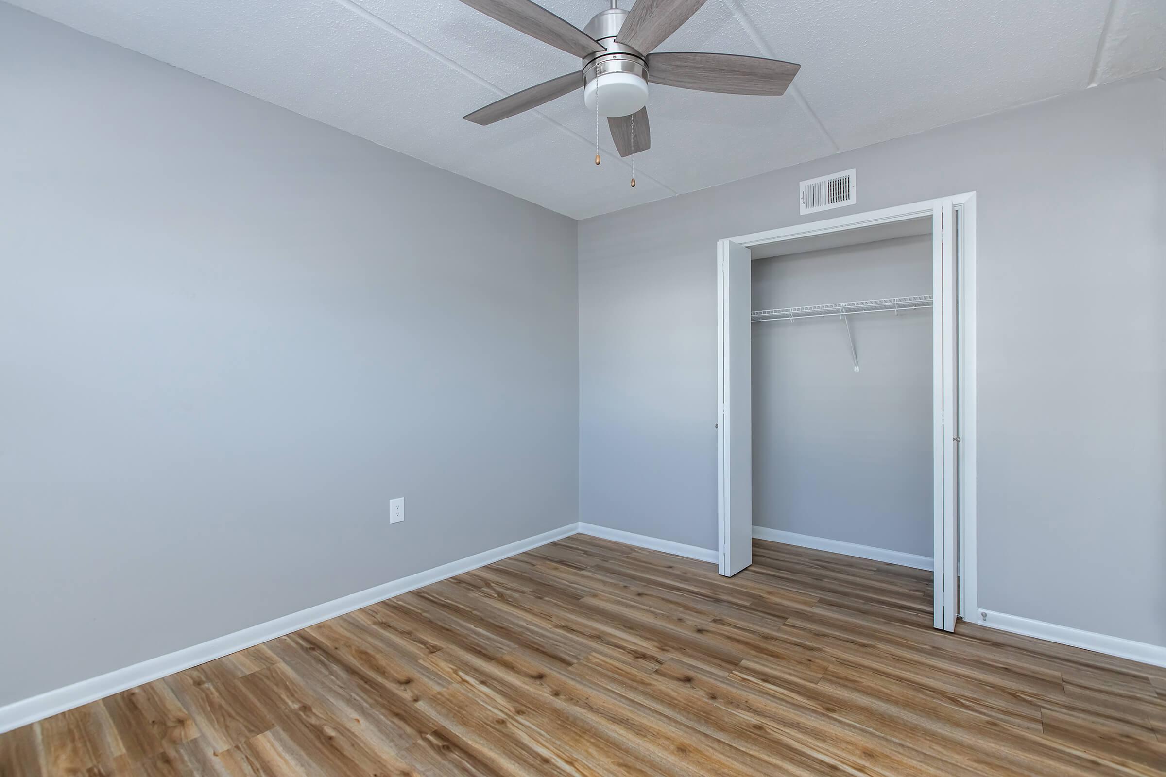A minimalist room featuring light gray walls and a ceiling fan. The floor is made of wood-like laminate. There is a closet with a white wire shelf and a door leading to it. The overall ambiance is bright and modern, with plenty of natural light.