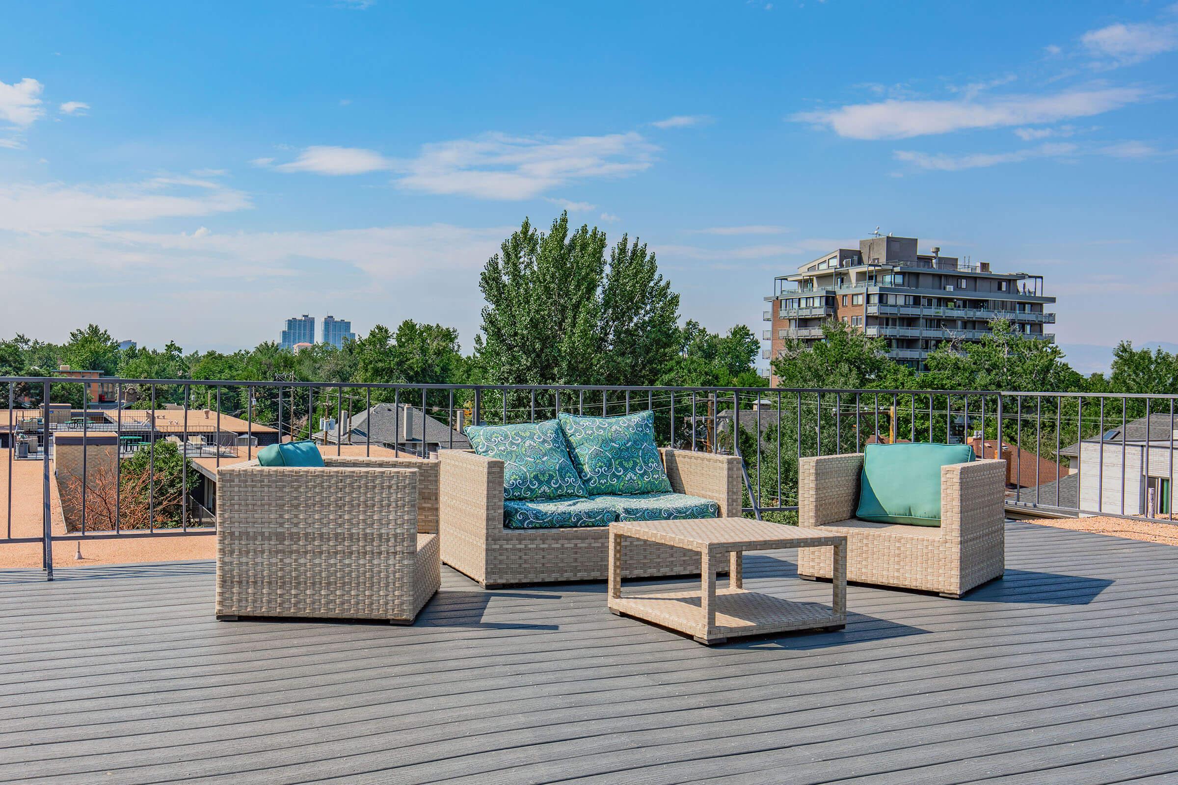 A rooftop patio featuring a light-colored wicker seating area with teal-patterned cushions. There is a small wicker coffee table in front of a couch and two armchairs. The scene is surrounded by trees and residential buildings under a clear blue sky.