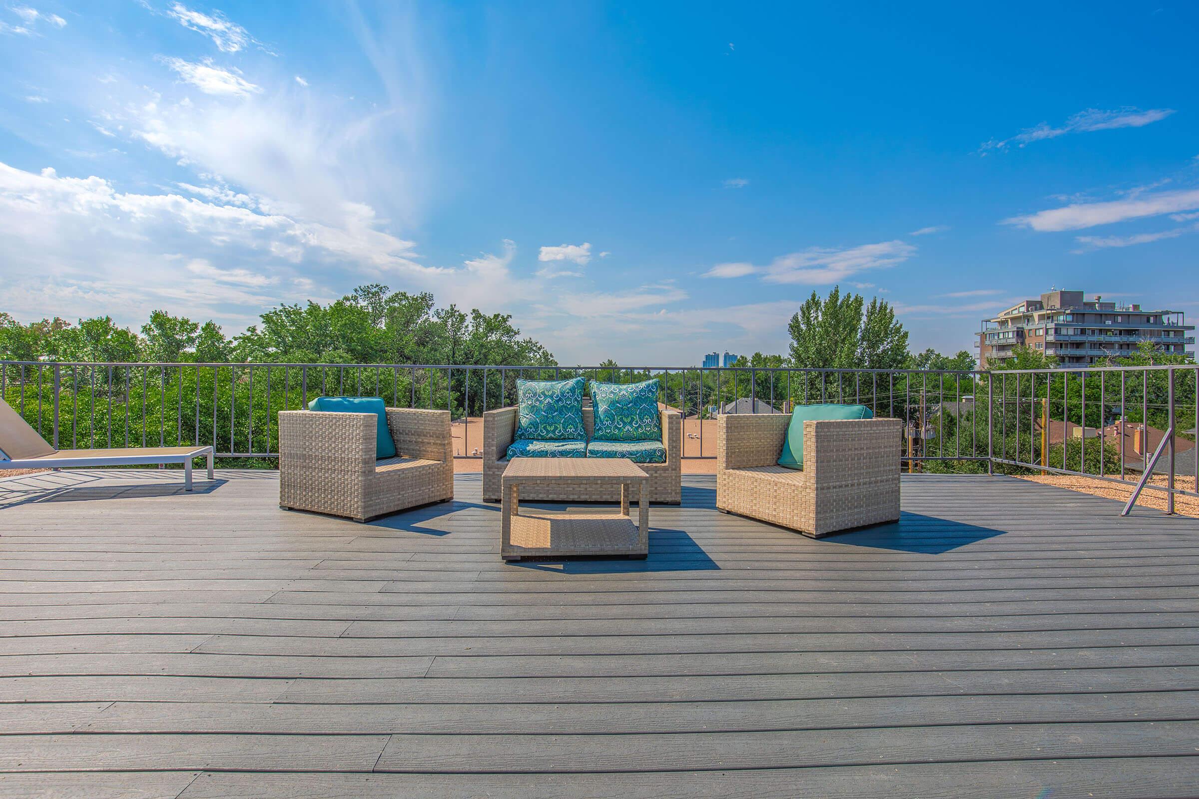 A rooftop terrace featuring modern wicker furniture with teal cushions, including a small table and two chairs. The space overlooks greenery and distant buildings under a clear blue sky.
