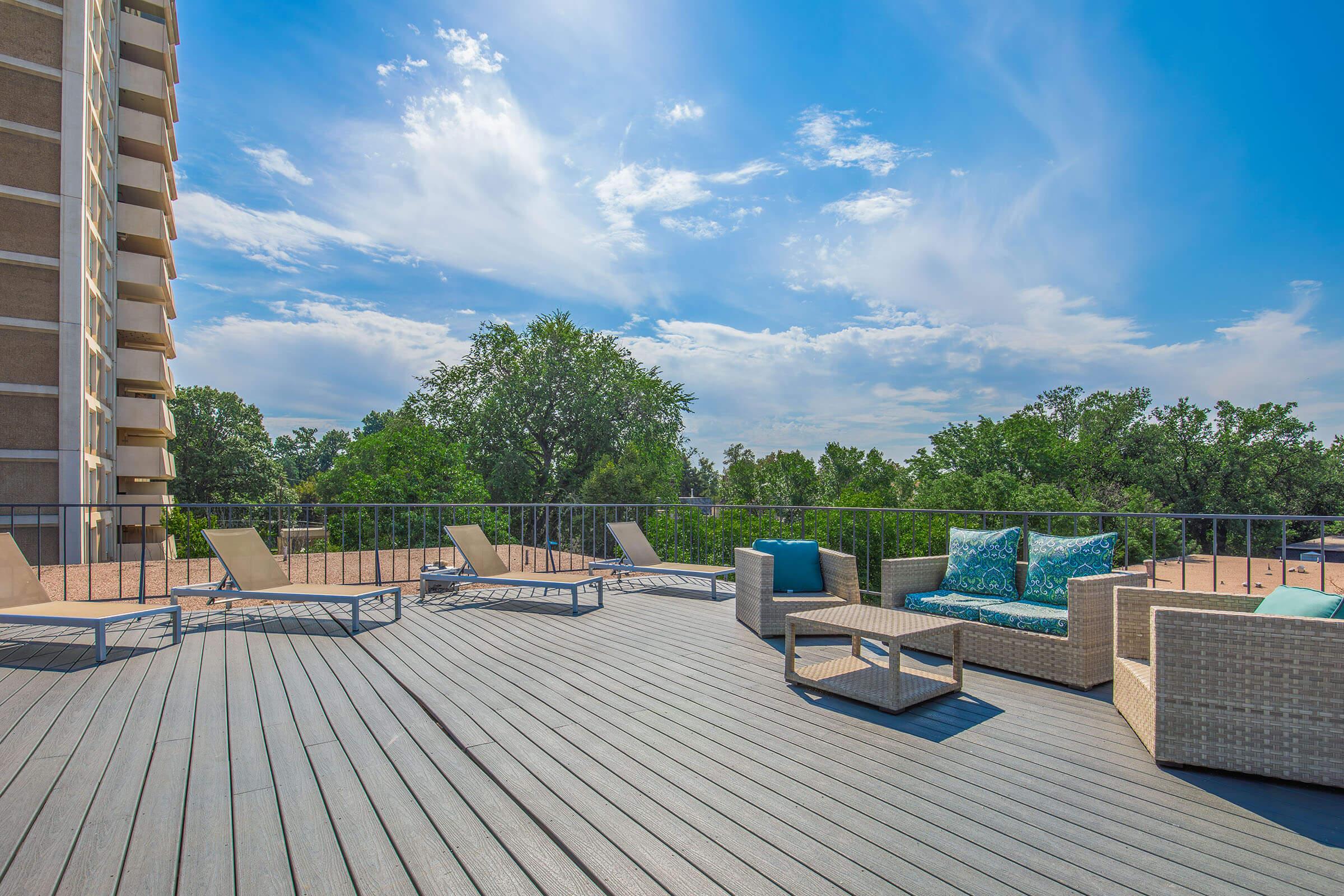 A sunny outdoor deck area featuring stylish lounge chairs and a seating arrangement with cushions. Surrounding greenery and a clear blue sky create a serene atmosphere, while a modern building stands nearby. Perfect for relaxation or social gatherings.