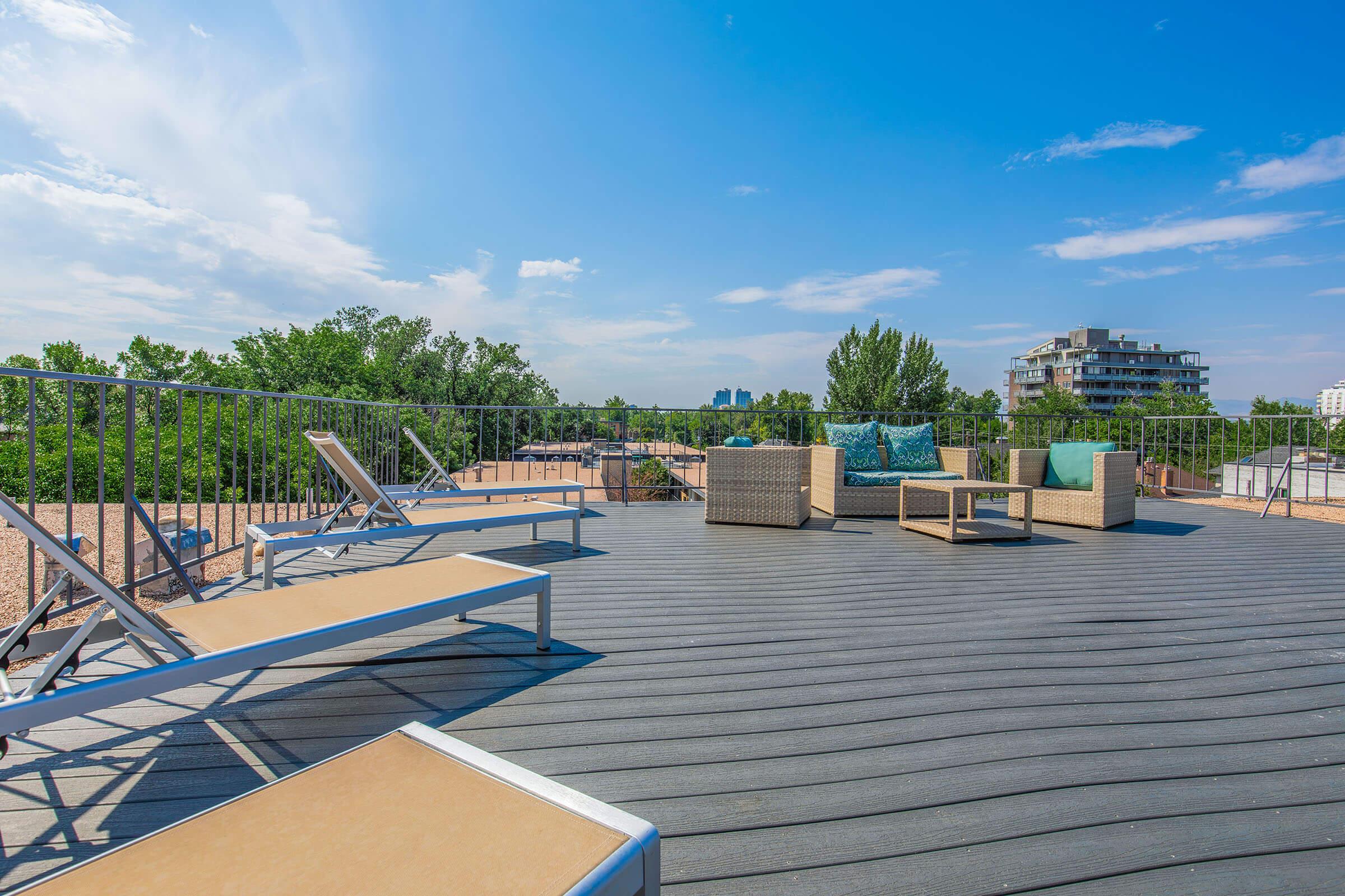 A spacious rooftop terrace featuring lounge chairs and a seating area with cushions, surrounded by greenery and blue sky. Modern buildings are visible in the background, creating a relaxed outdoor atmosphere.