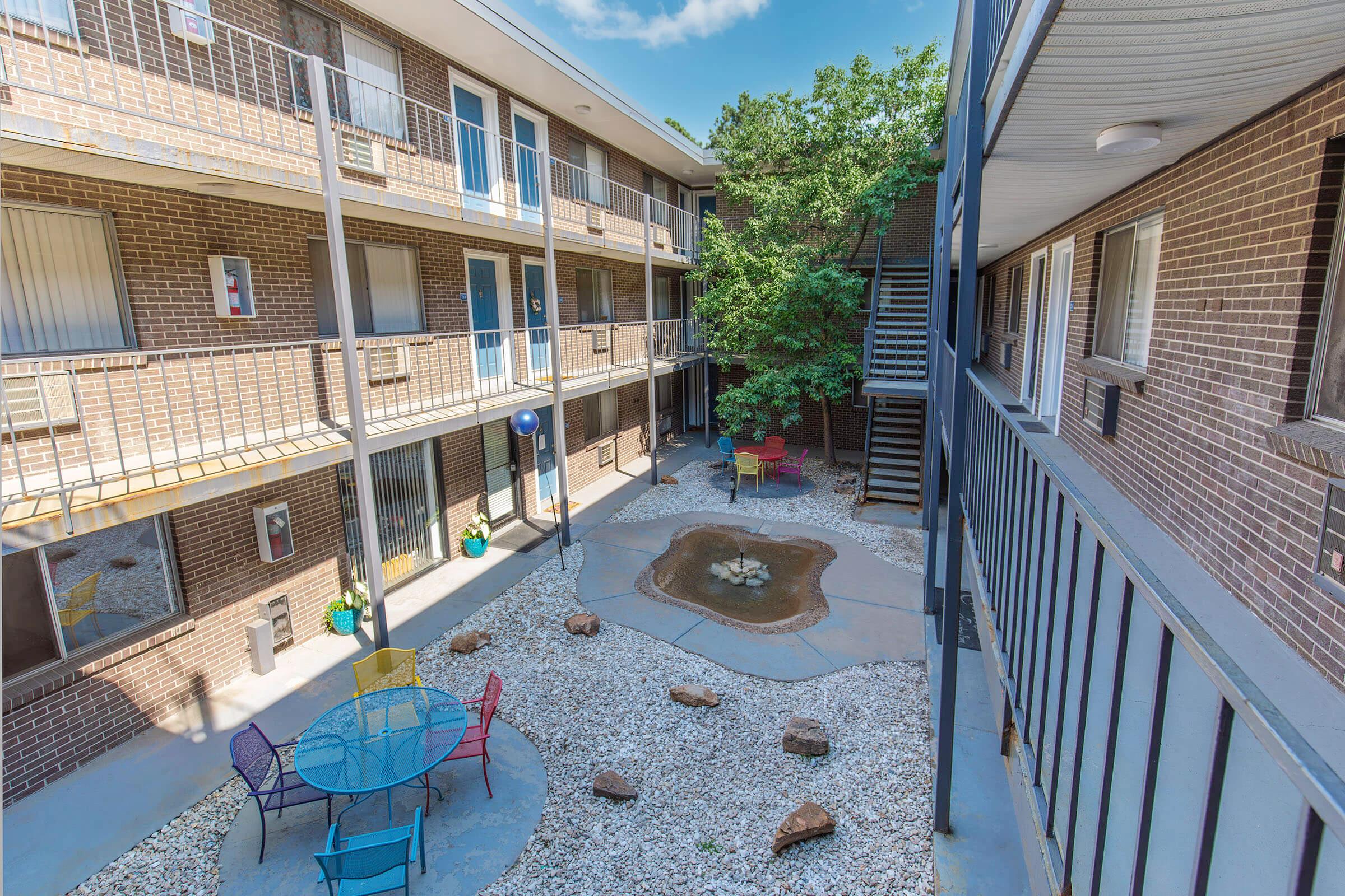 View of an outdoor courtyard in an apartment complex, featuring colorful seating around a circular table, small rock formations, a water feature, and surrounding greenery. The buildings are two stories high with balconies overlooking the courtyard. Natural light brightens the area, creating an inviting atmosphere.