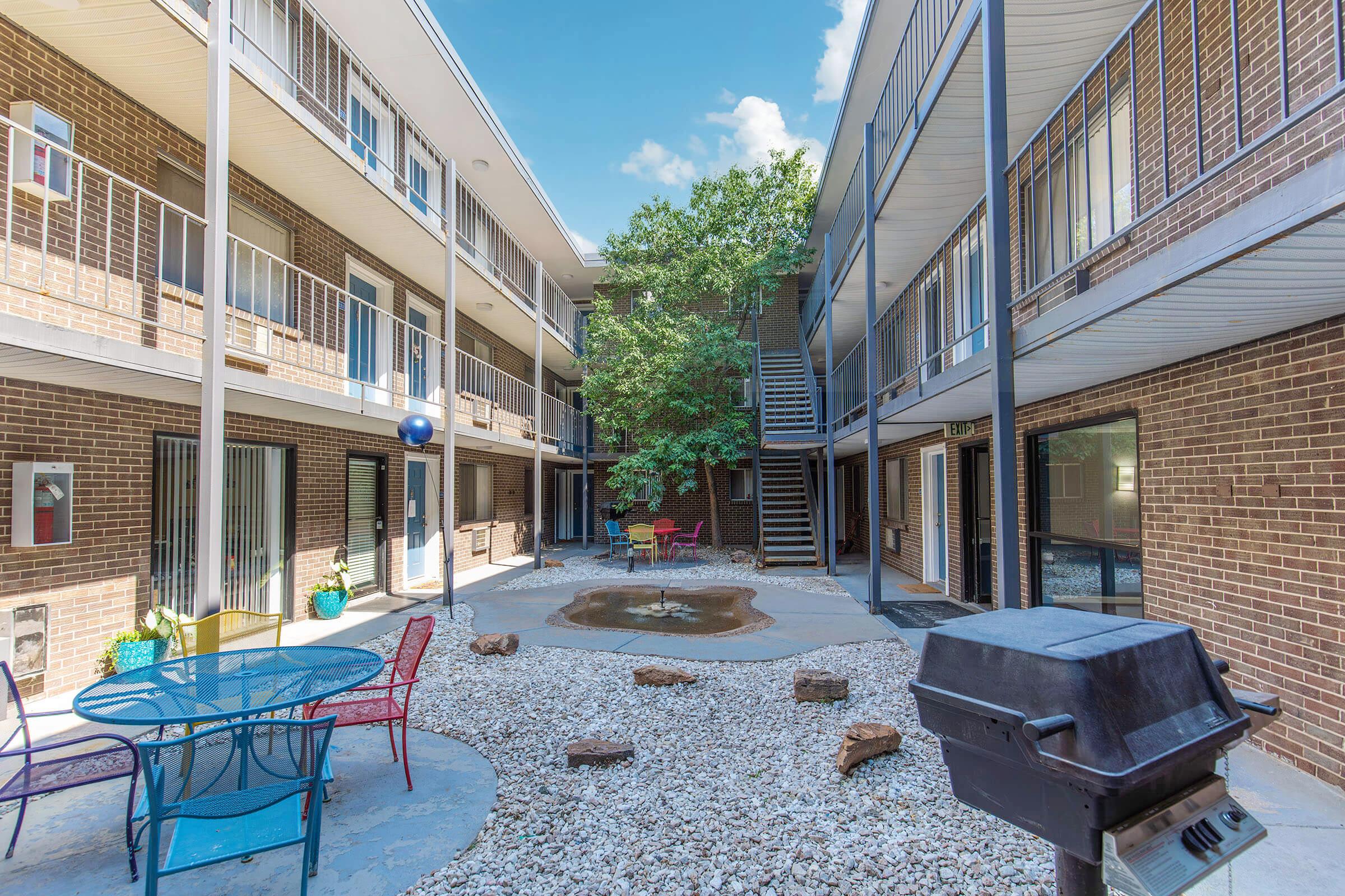 A courtyard of an apartment complex featuring a small pond surrounded by rocks, a circular table with chairs, and a grill. The area is landscaped with a tree and has a modern design, providing a relaxing outdoor space. Nearby balconies overlook the courtyard.