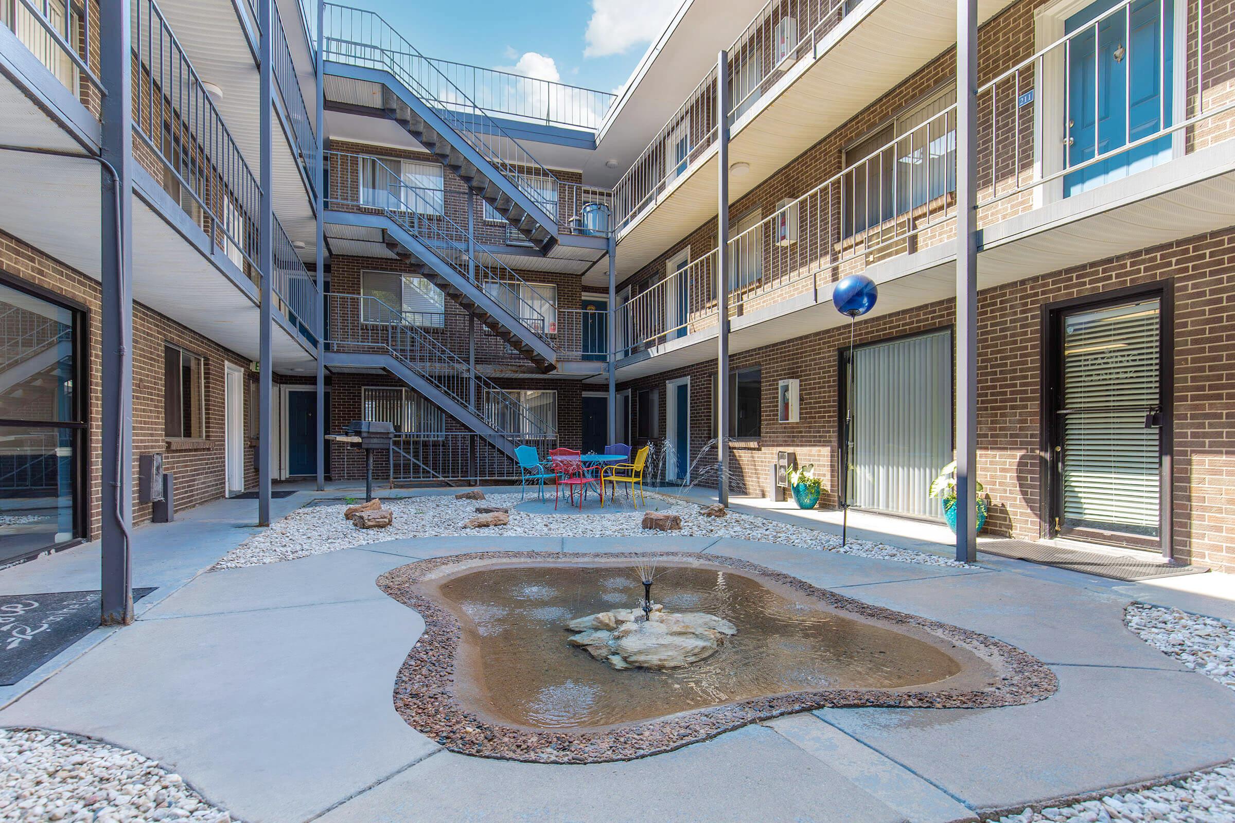 A courtyard view of an apartment building featuring a central stone fountain surrounded by gravel. The courtyard is bordered by multiple levels of balconies, with outdoor seating in vibrant colors. Stairs lead to upper levels, and there are potted plants along the ground floor. Bright blue sky overhead.