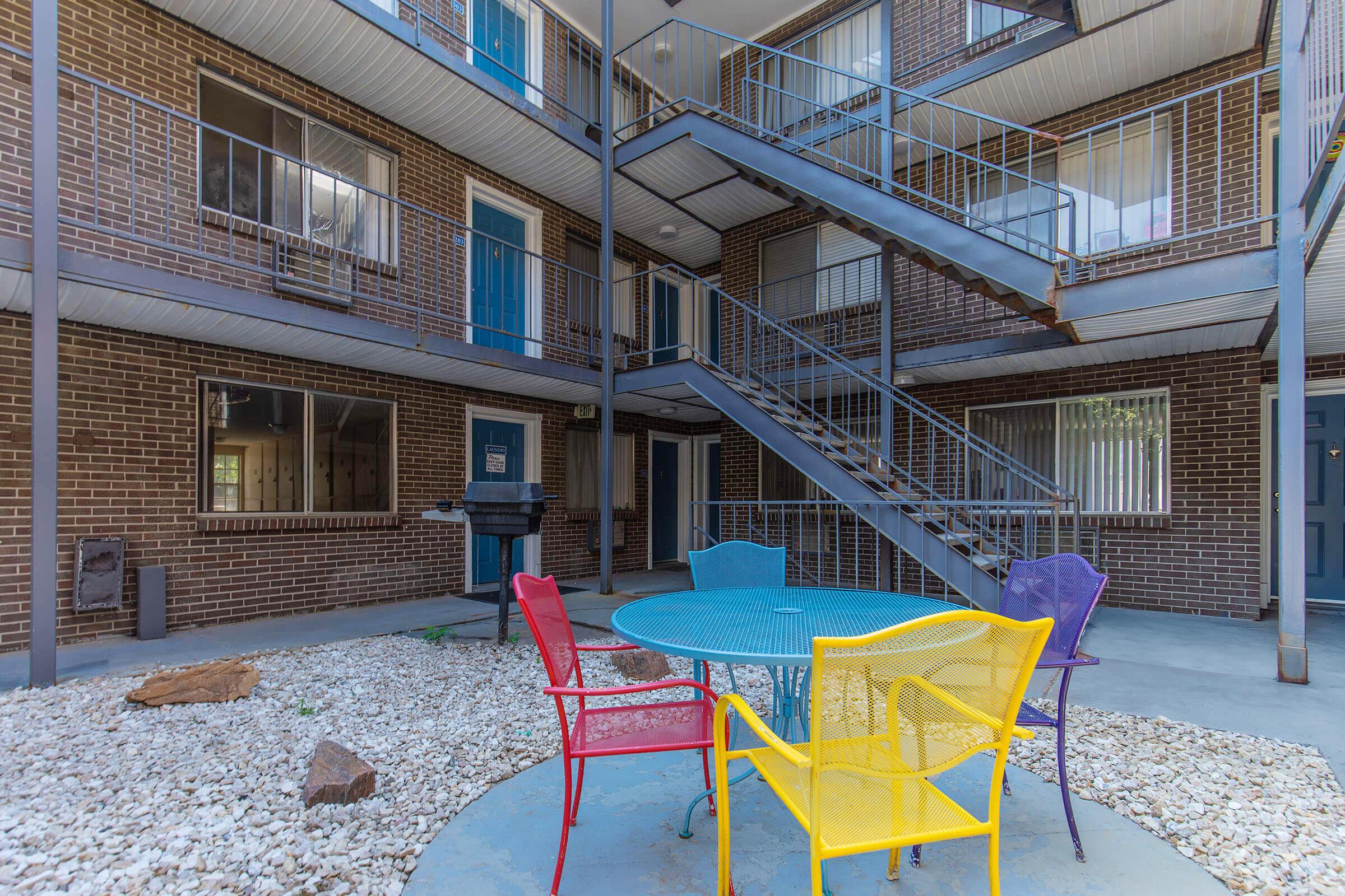 A cozy courtyard area featuring a round table surrounded by colorful chairs in red, yellow, and purple. In the background, there's a multi-level building with a metal staircase and several windows, set against a backdrop of gravel and a small rock.