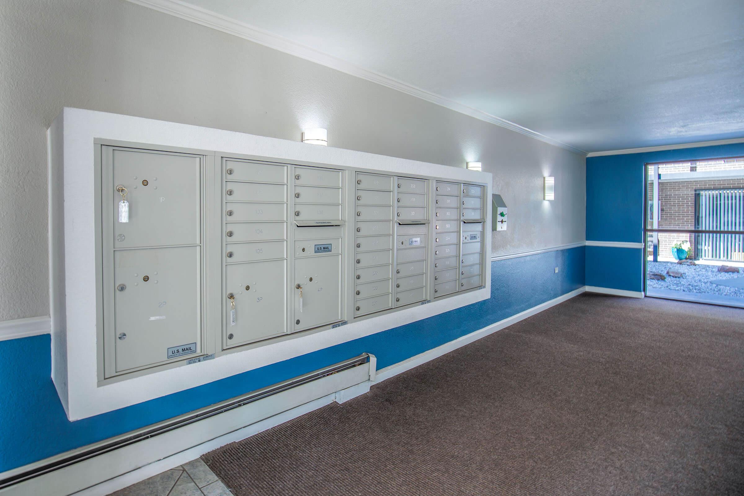 Mailboxes mounted on a wall in a well-lit hallway with blue and white walls. The floor is carpeted in a neutral color, and there are two wall-mounted lights providing illumination. The area appears clean and organized, suitable for residential or office mail delivery.