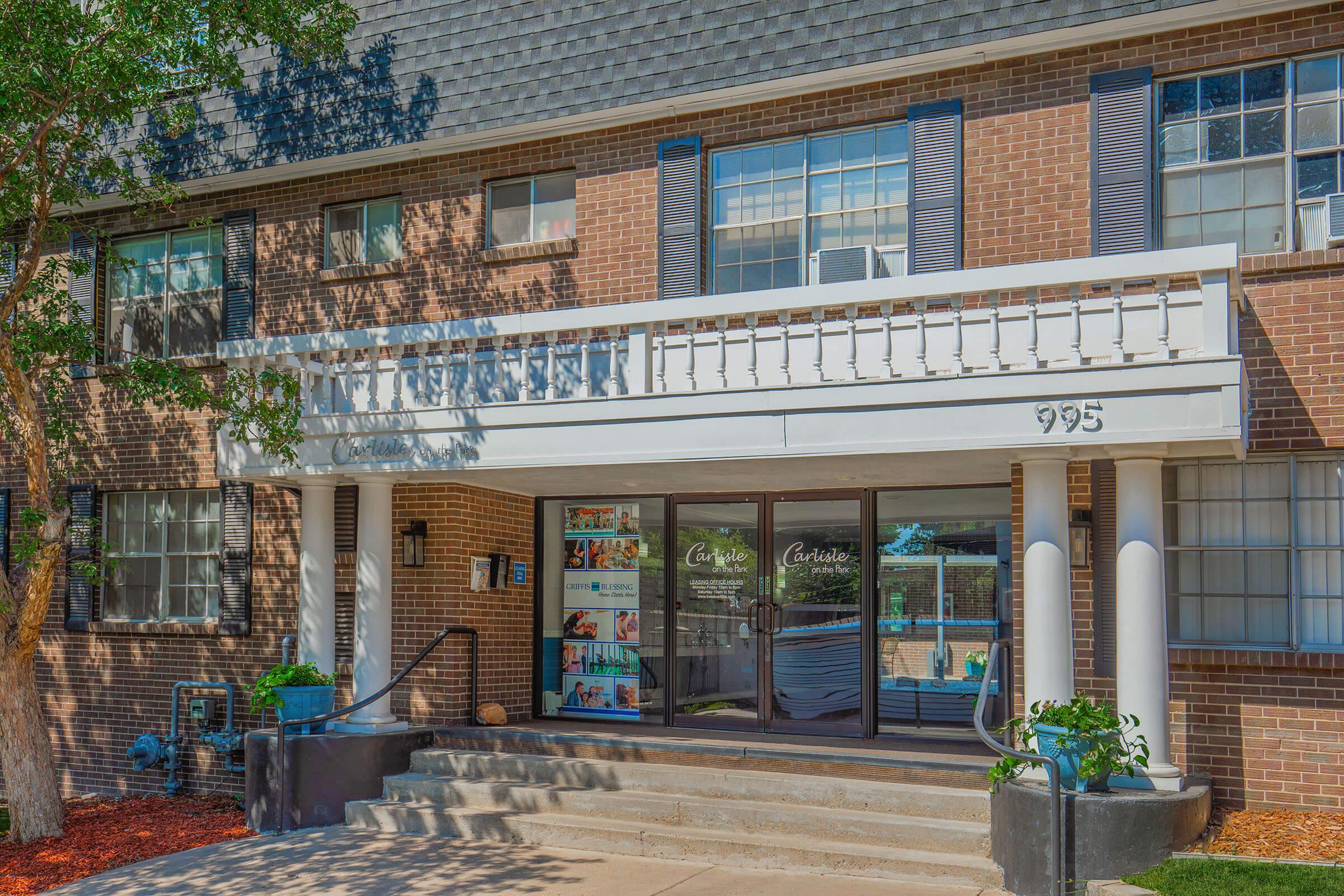 The image shows the entrance of a brick building with a white awning displaying the number "995." Flanking the entrance are large windows with dark frames. To the side, there are promotional displays visible through the glass. A tree and landscaped area are present in front of the building.