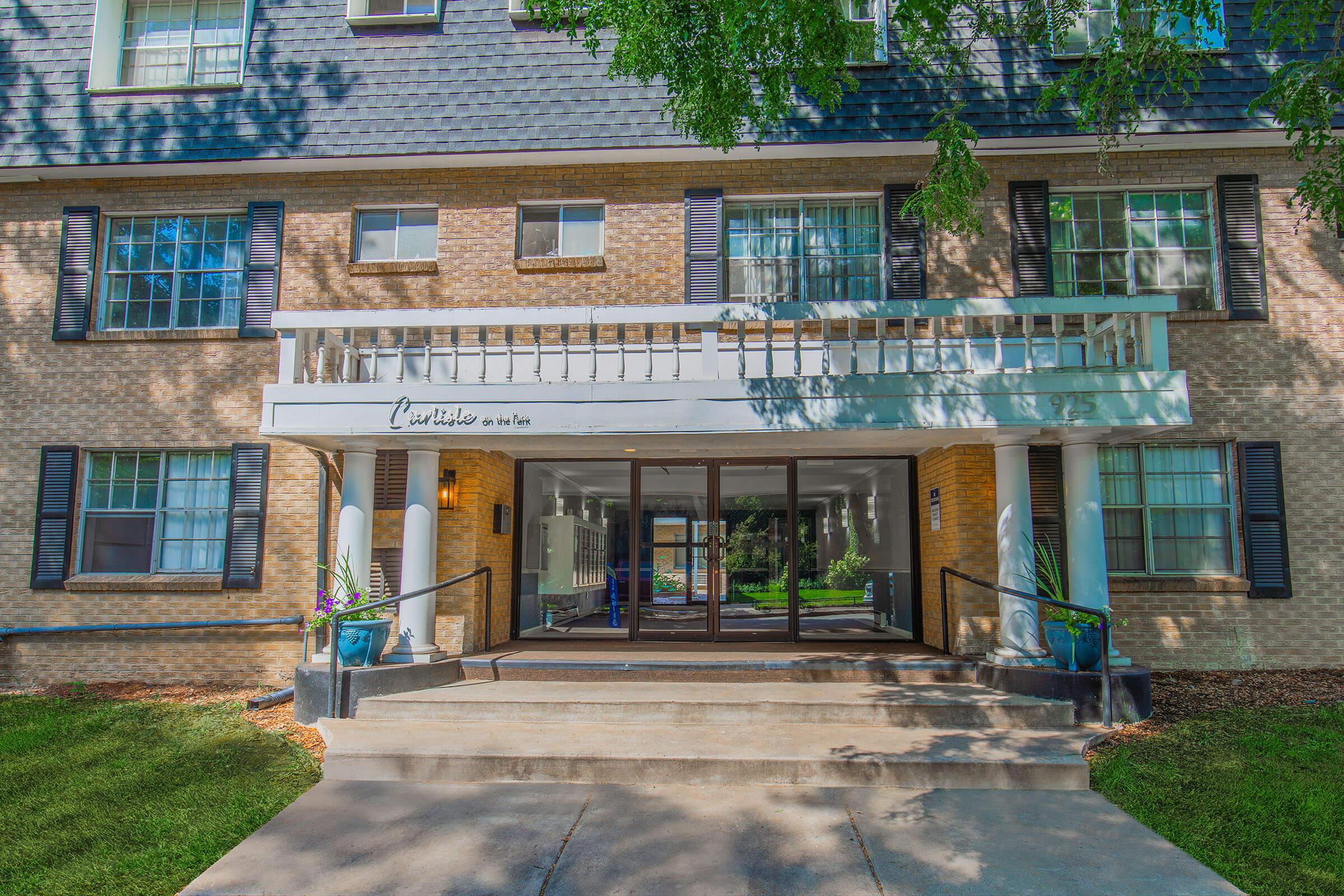 Entrance of a multi-story building with a brick facade and large windows. The entrance features sliding glass doors and a covered porch with decorative columns. Potted plants are placed near the entrance, and greenery is visible in the background. The building is well-lit and inviting.